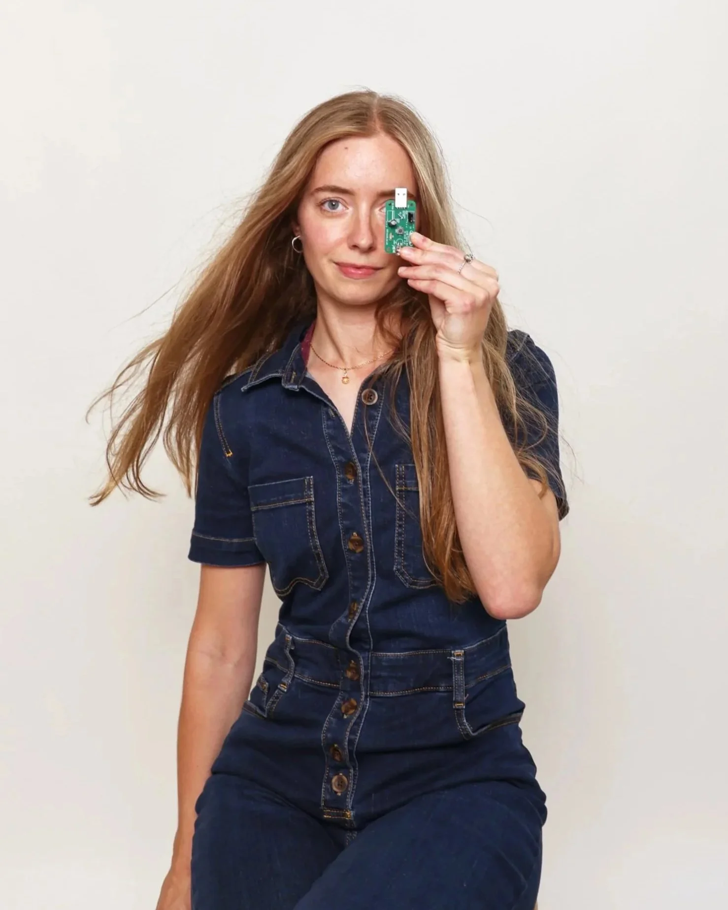 A young woman with long, wavy red hair, wearing a dark denim button-up shirt with short sleeves, sitting against a plain white background. She is holding a small electronic circuit board up to her right eye, covering it, while her left eye is open, looking at the camera. She has a slight smile, small hoop earrings, a delicate necklace, and rings on her fingers.
