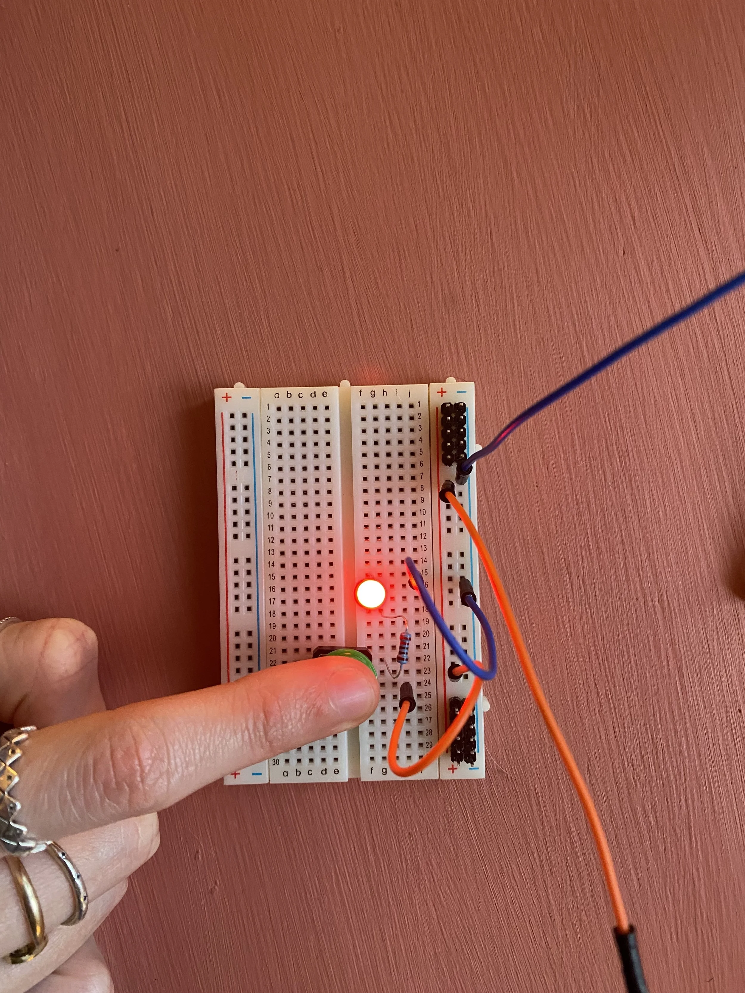 Breadboard with LED lamp growing and finger pressing down on switch on pink background