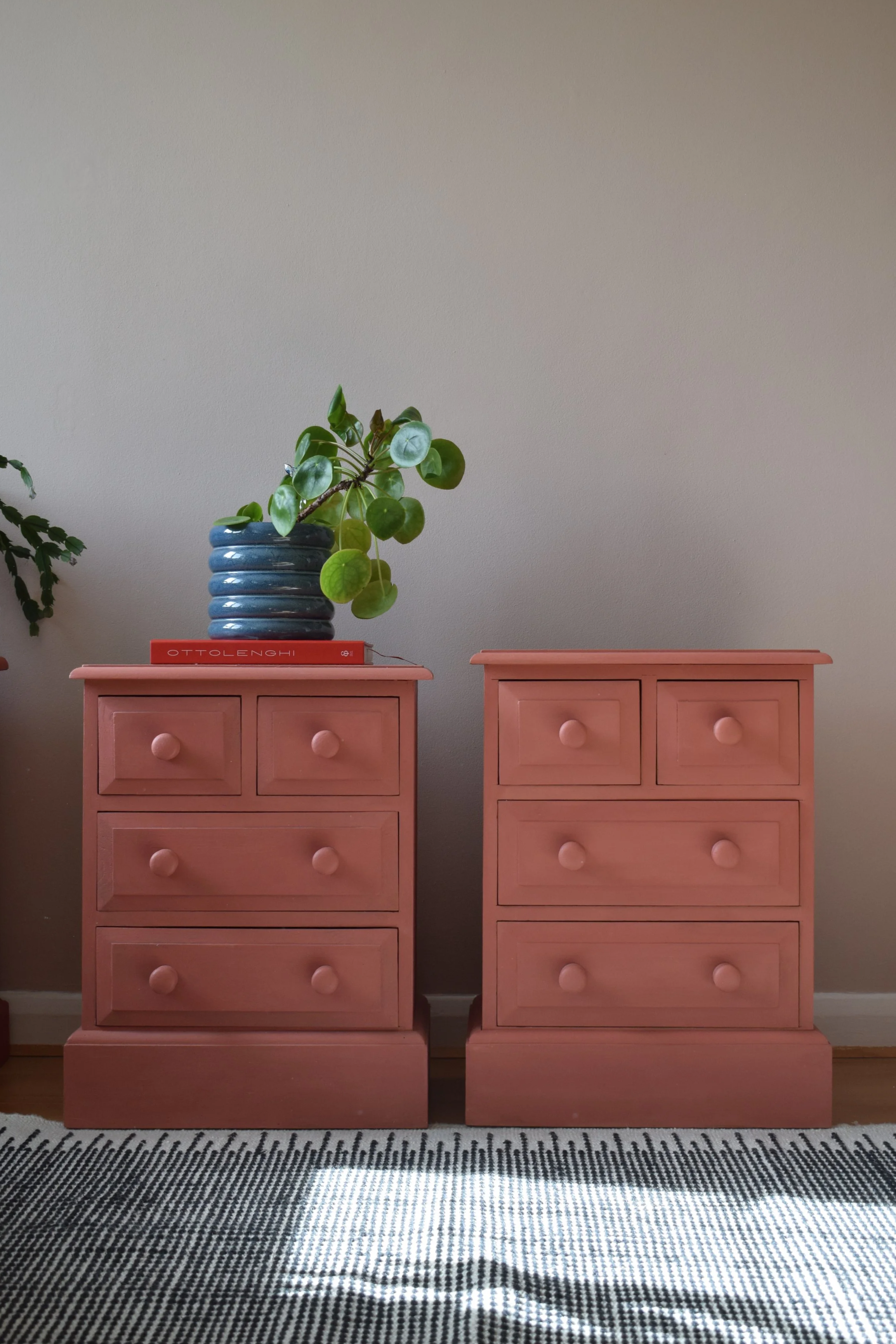 Two pink wooden dressers with round knobs, each with three drawers, placed side by side against a beige wall. The left dresser has a blue ribbed vase with a green plant on top, and the right dresser has a closed pink box. Both dressers are on a patterned black-and-white rug.