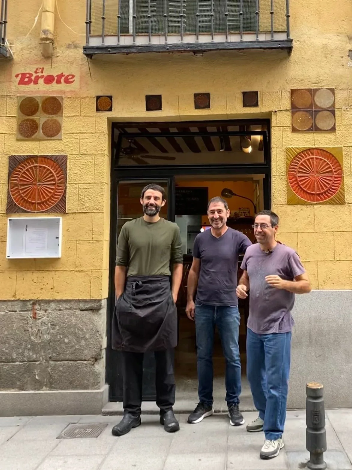 Tres hombres sonriendo frente a un restaurante con fachada amarilla y decoraciones de manera circular y cuadrada en la pared, uno con delantal negro, y los otros dos con camisetas moradas y pantalones jeans.