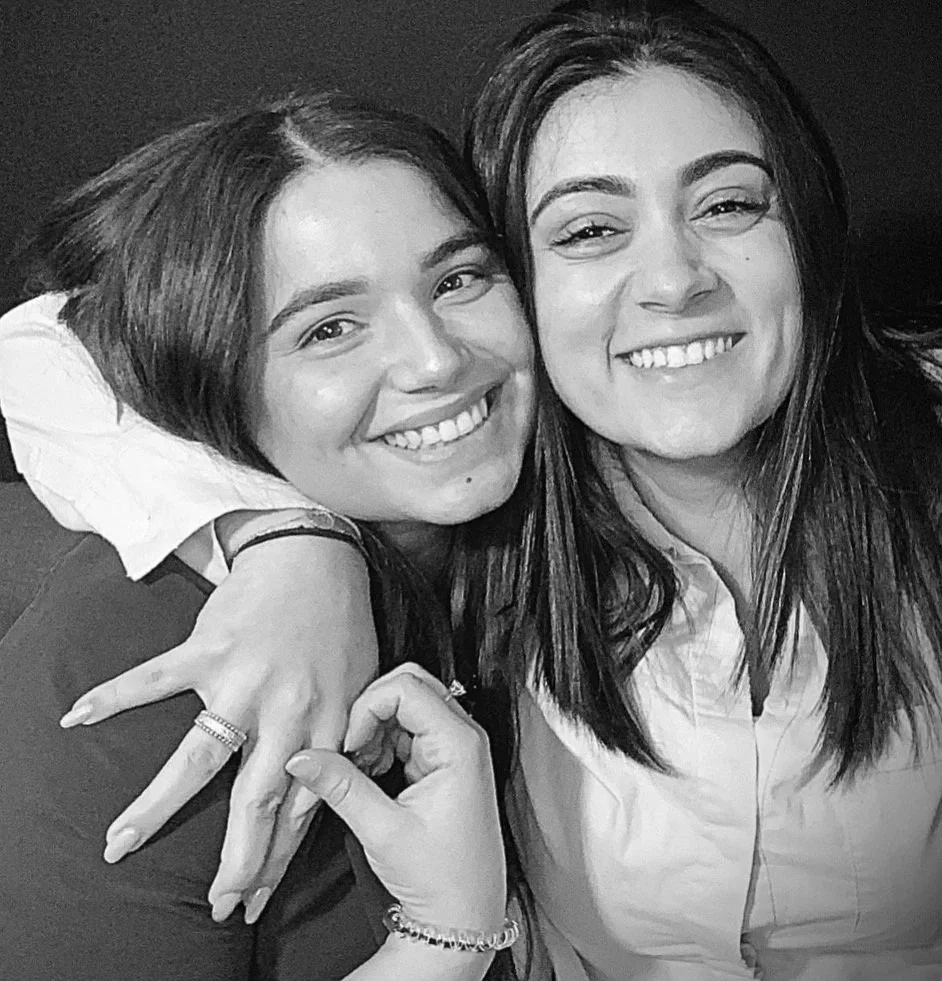 Deux femmes souriantes posent pour une photo en noir et blanc, l'une d'elles est en train d'embrasser l'autre sur la joue, toutes deux ont de longs cheveux. La femme à gauche porte une veste noire et un bracelet, la femme à droite porte une chemise claire.