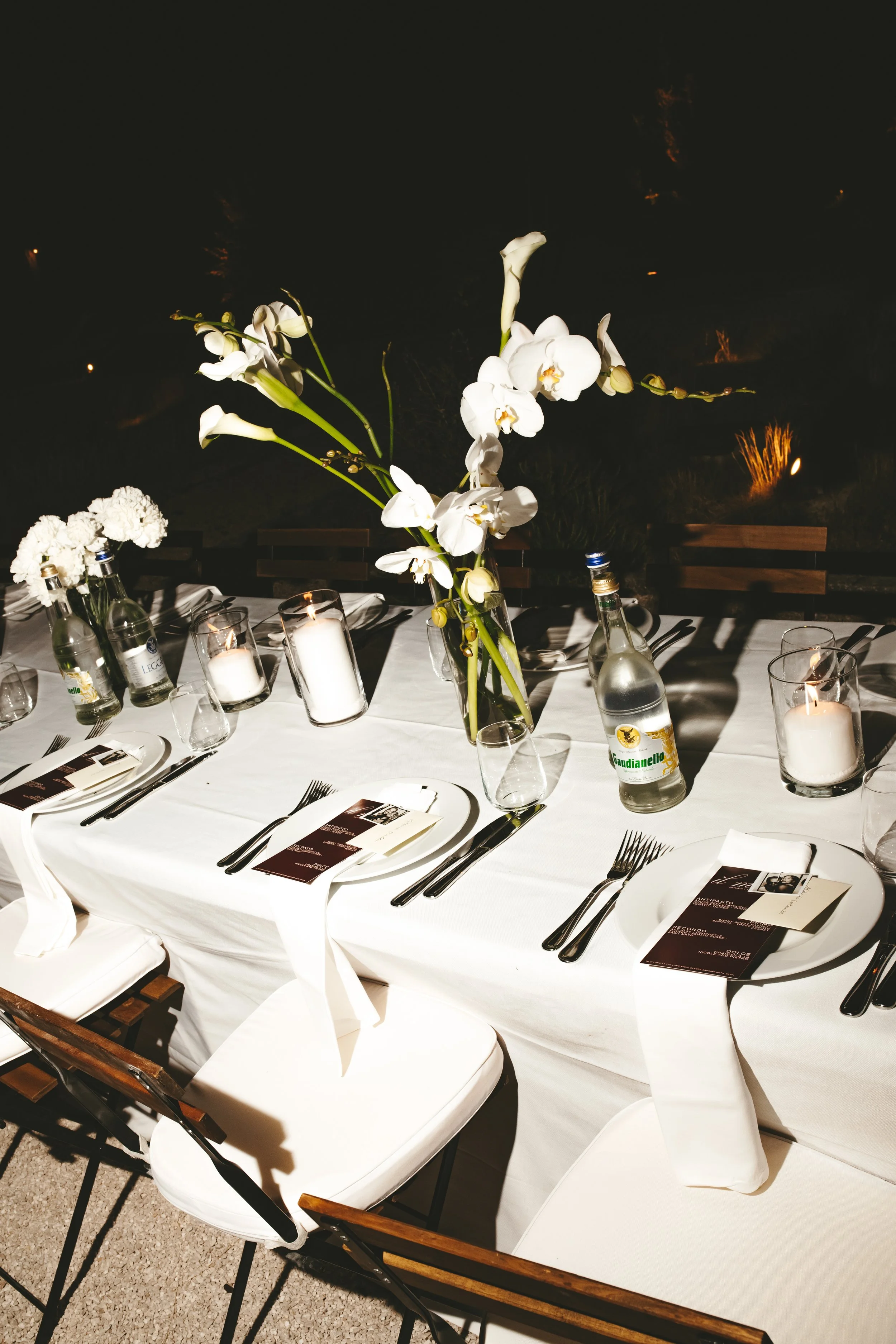 A table set for a formal dinner with white tablecloths, black knives, forks, and glasses. A floral centerpiece features white orchids and carnations, and there are lit candles in glass holders. Menus or programs are placed on each plate.