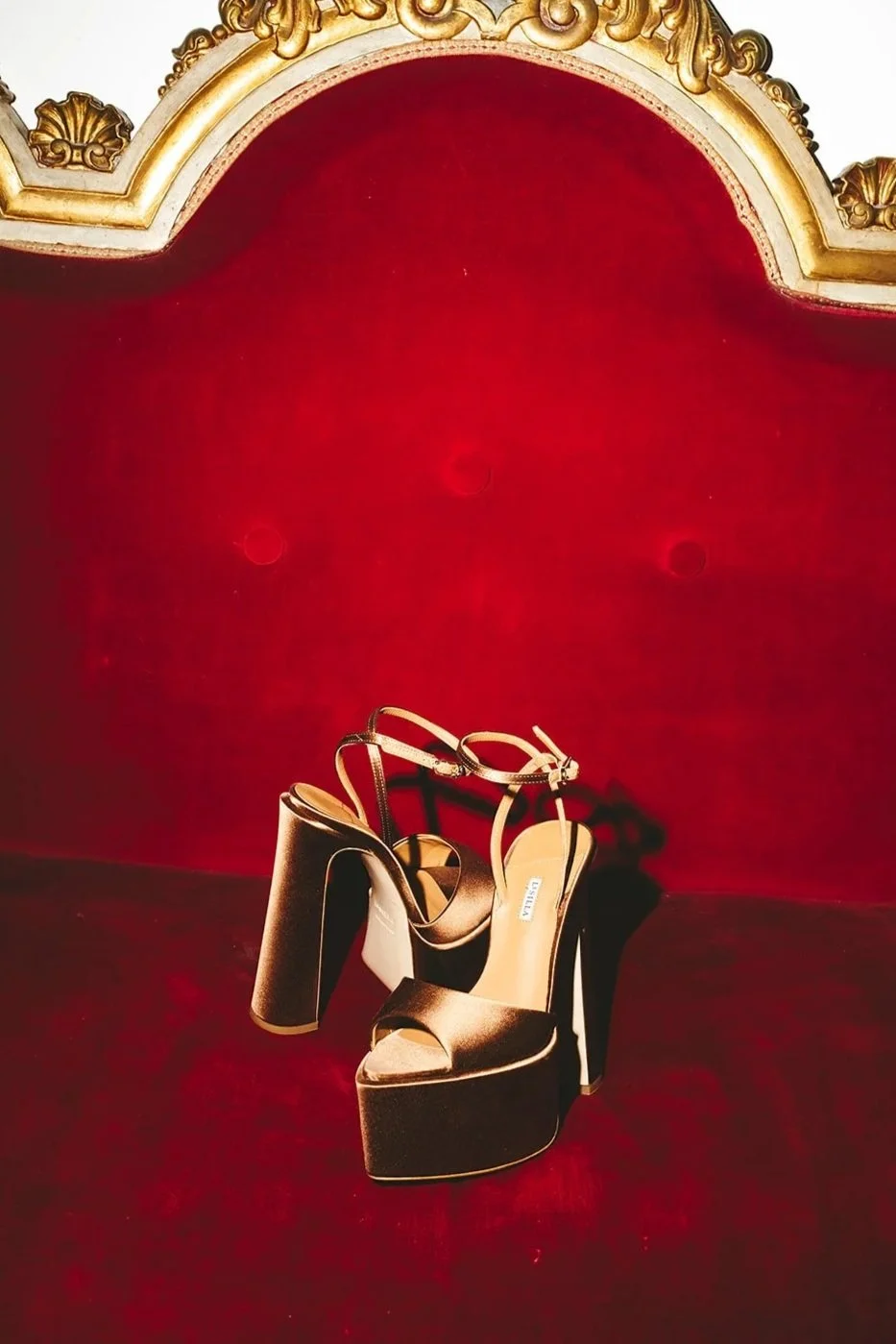 Pair of gold high-heeled platform shoes with ankle straps on a red velvet surface, with a red velvet and gold ornate backdrop.