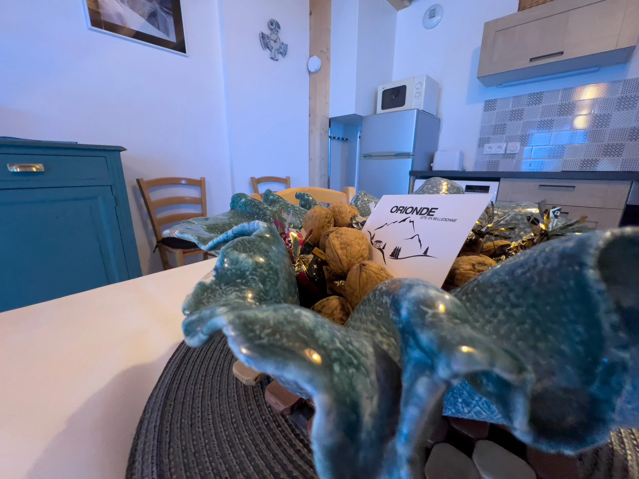 A table with wrapped food items and a card labeled 'Orionde Gîte en Bellecombe' in a room with blue lighting, wooden chairs, a kitchen area with a microwave and fridge, and wall decorations including a framed picture and a decorative fish.