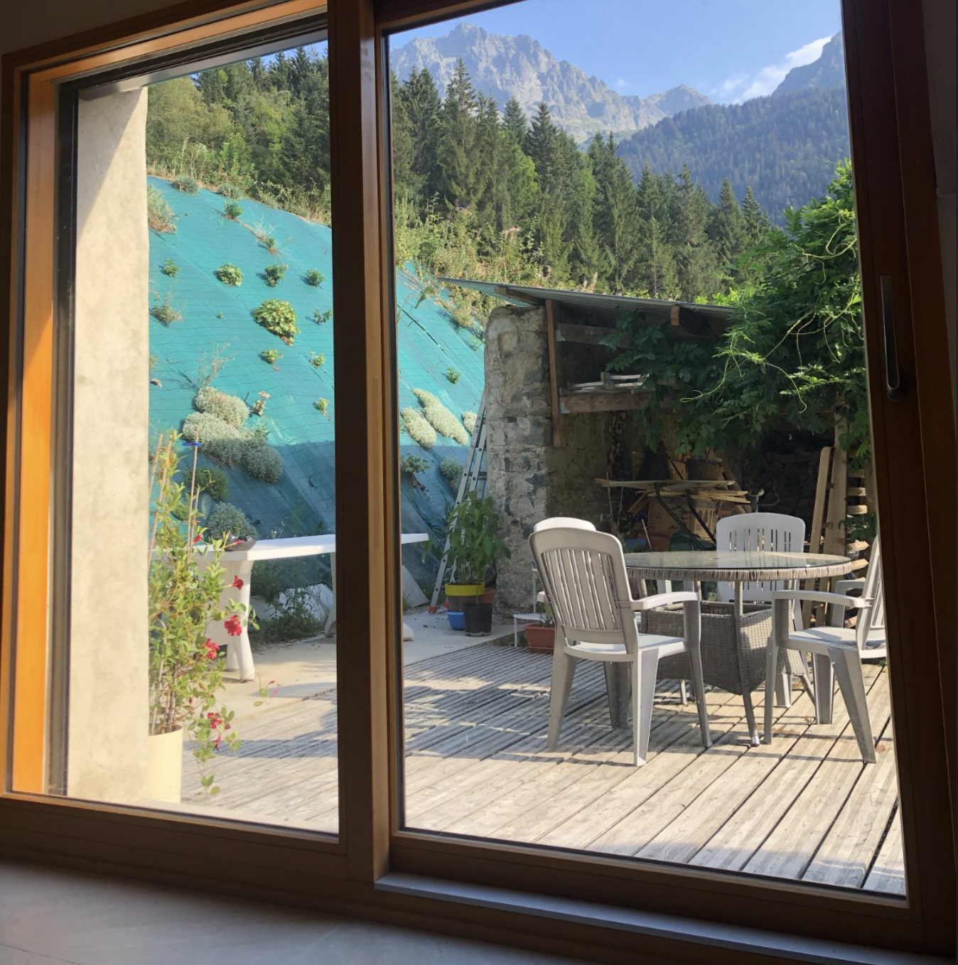 View through a glass door showing an outdoor patio with a round table and four white chairs, surrounded by potted plants, a stone wall, and a lush green mountainous landscape in the background.