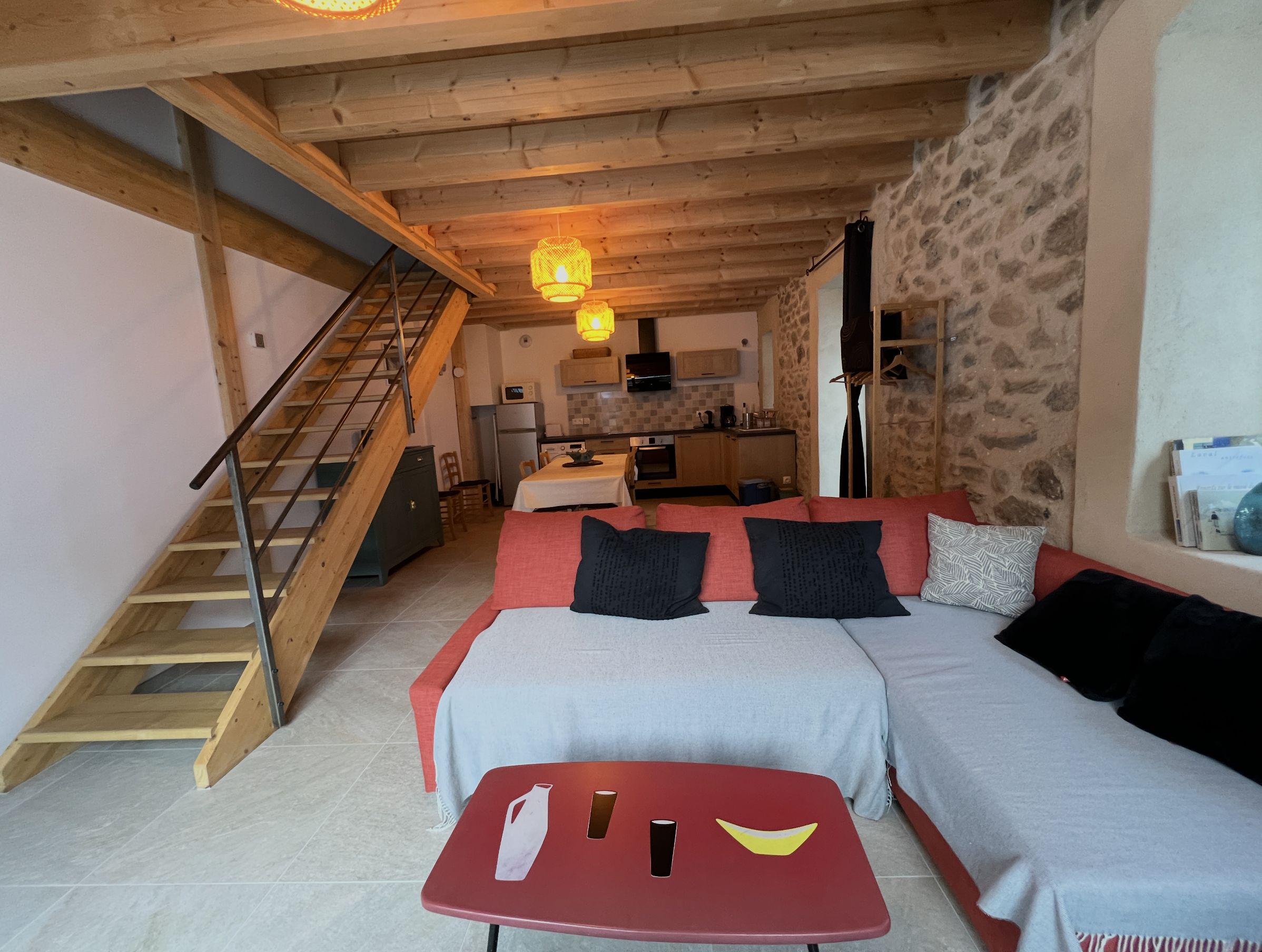 Living room with wooden ceiling, hanging orange lights, stone wall, and red sofa with multiple black and beige pillows. There is a small table with a game box in the foreground and a dining table in the background, along with a kitchen area.