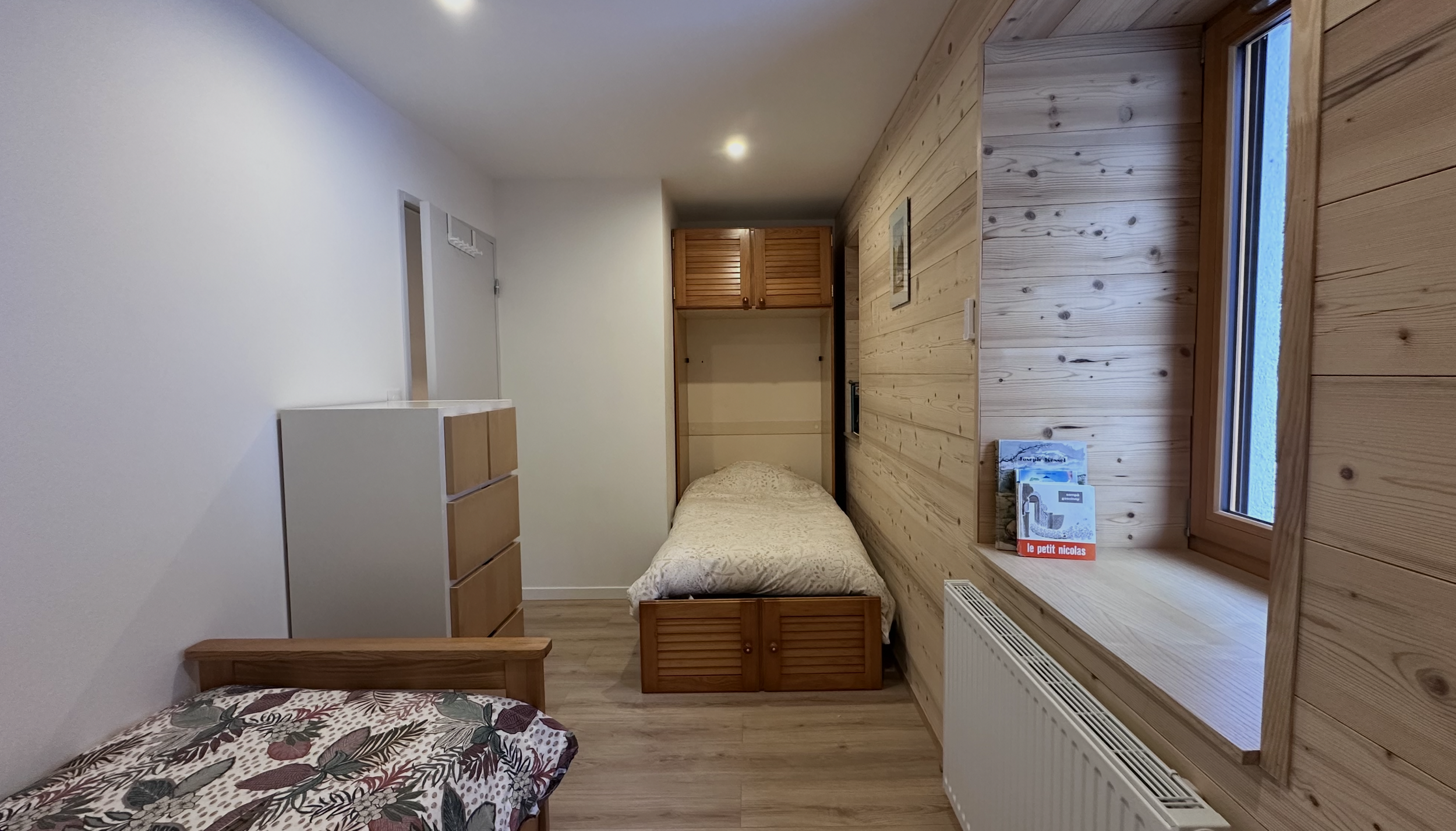 A cozy bedroom with two single beds, one decorated with a floral duvet, wooden furniture, a window with a panoramic view, and a wood-paneled wall.