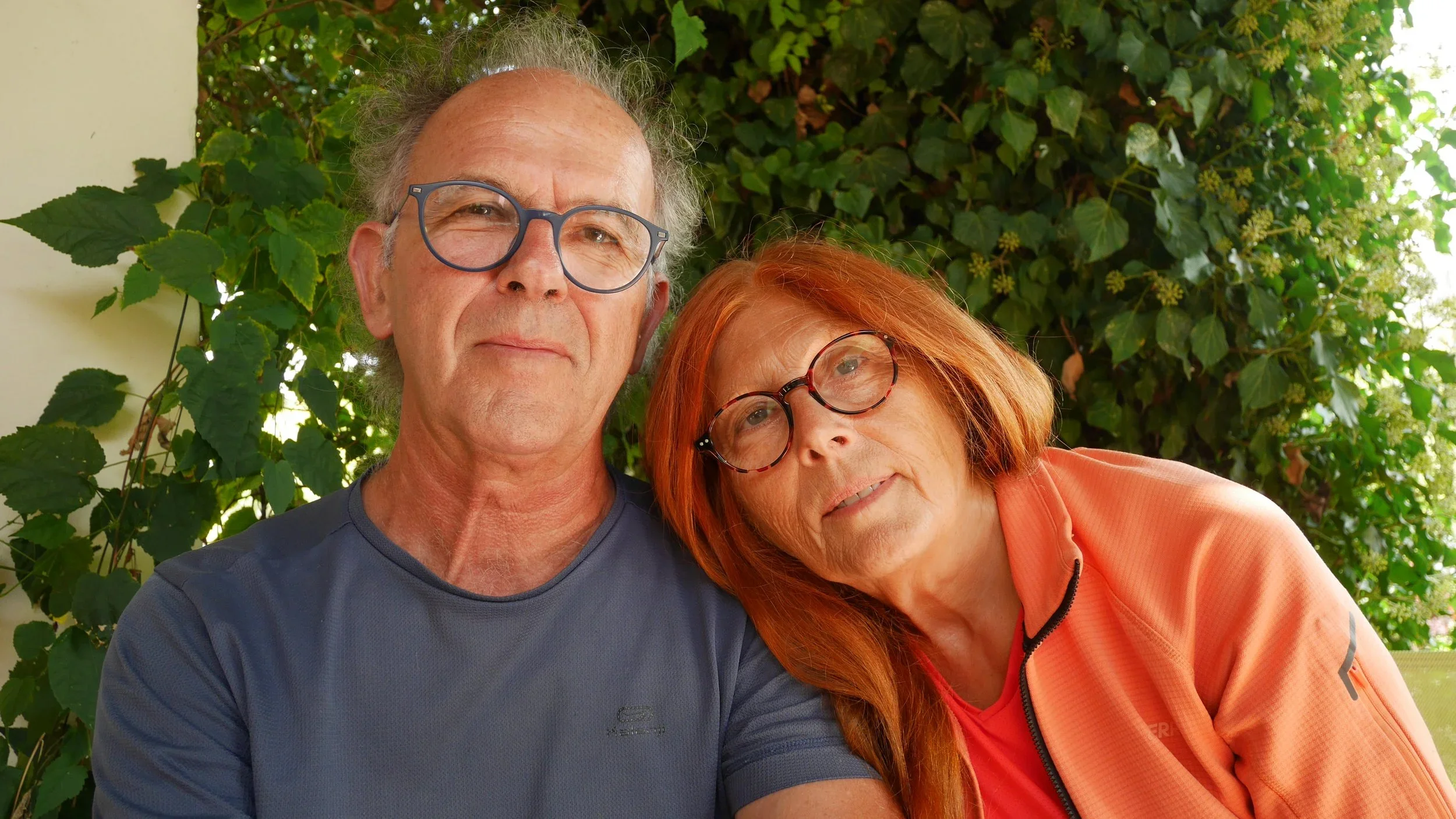 An elderly man and woman taking a selfie outdoors, with greenery and leaves in the background.