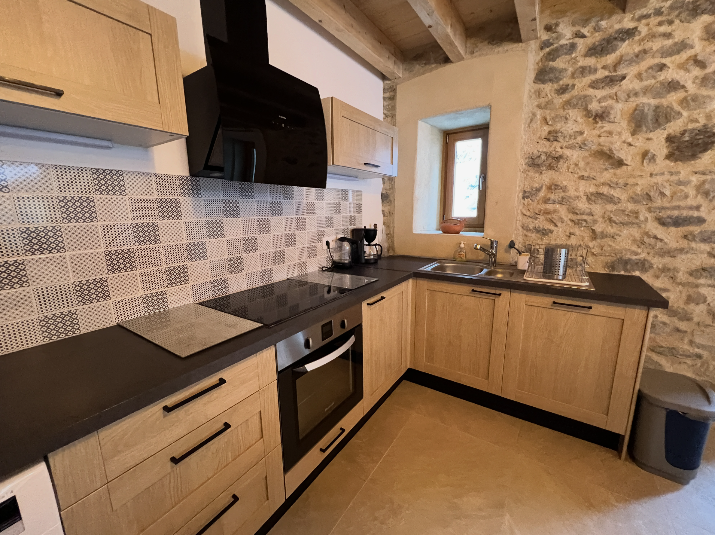 A kitchen with wooden cabinets, a black oven, patterned tile backsplash, small window, stone wall, and kitchen appliances on the countertop.