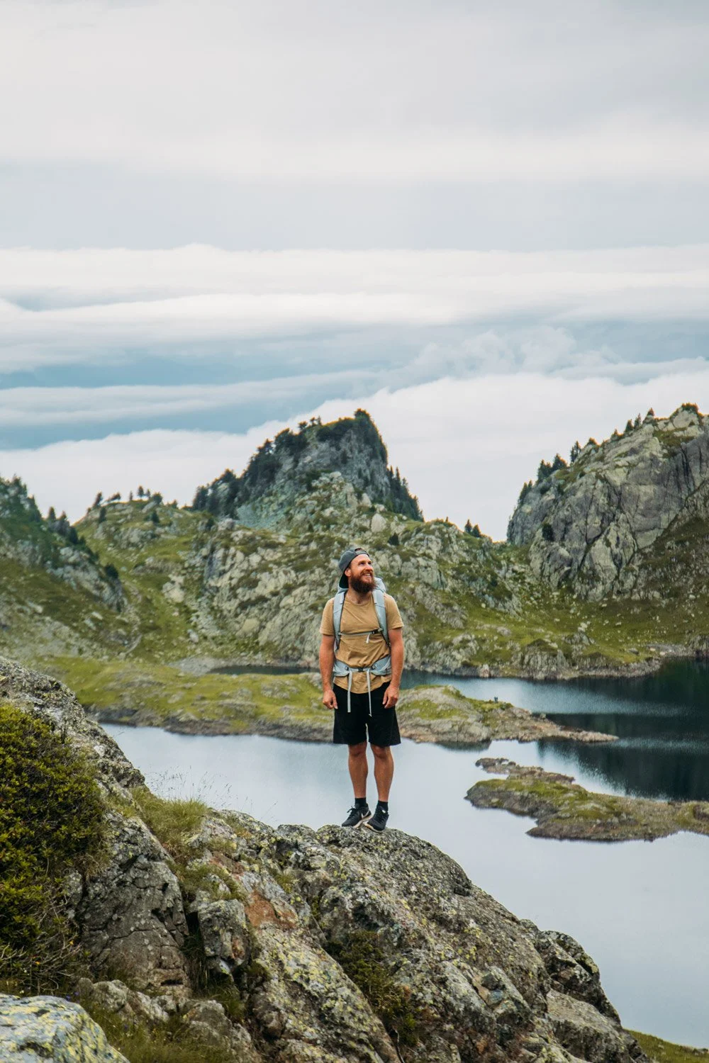A man with a beard wearing a backpack, shorts, and a cap stands on a rocky ledge above a body of water in a mountainous landscape with overcast skies.