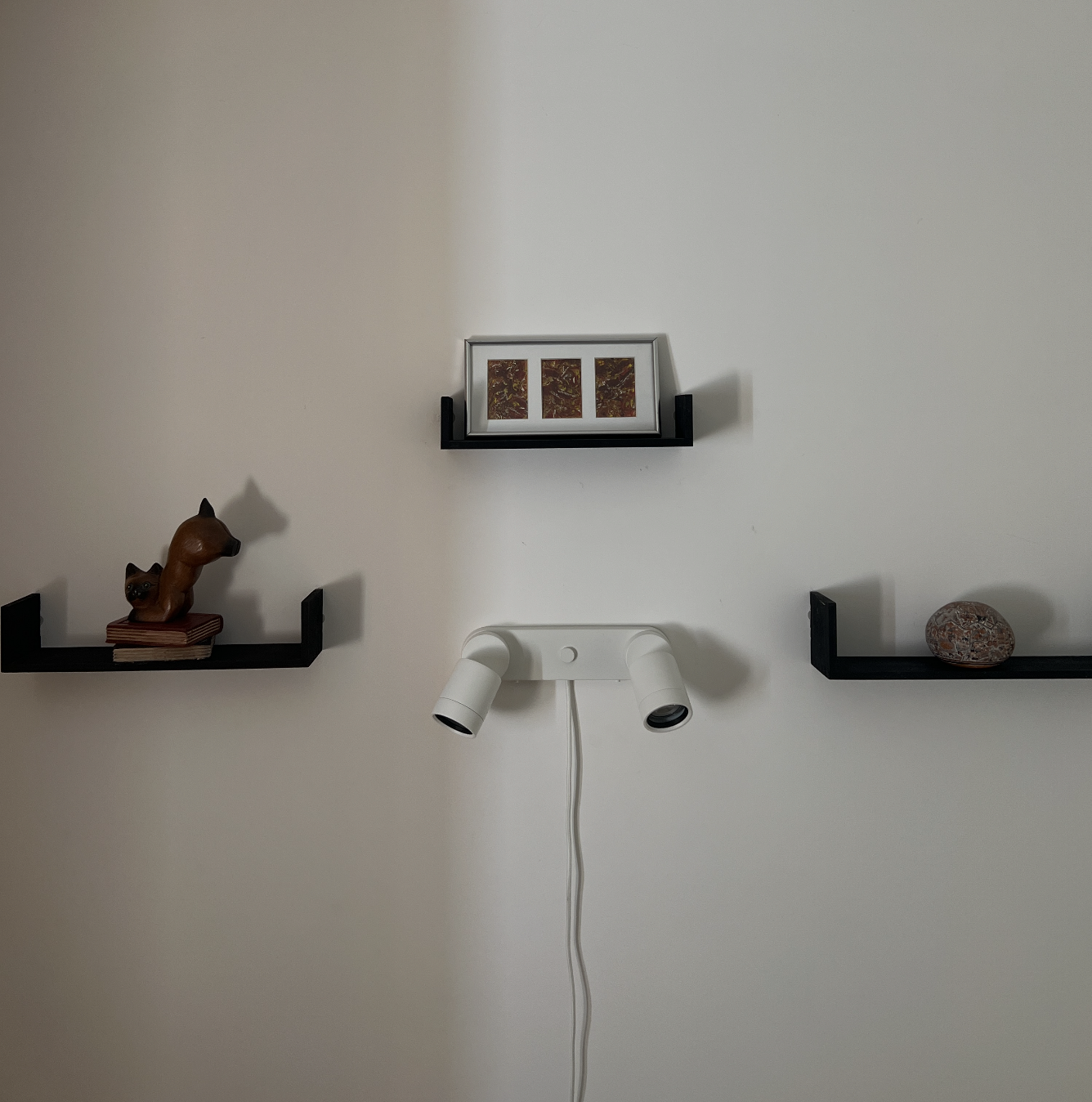 Wall with three black floating shelves, a framed picture, a small sculpture of a dog and cat on the left shelf, a stone on the right shelf, and a white ceiling light fixture with two adjustable spotlights.