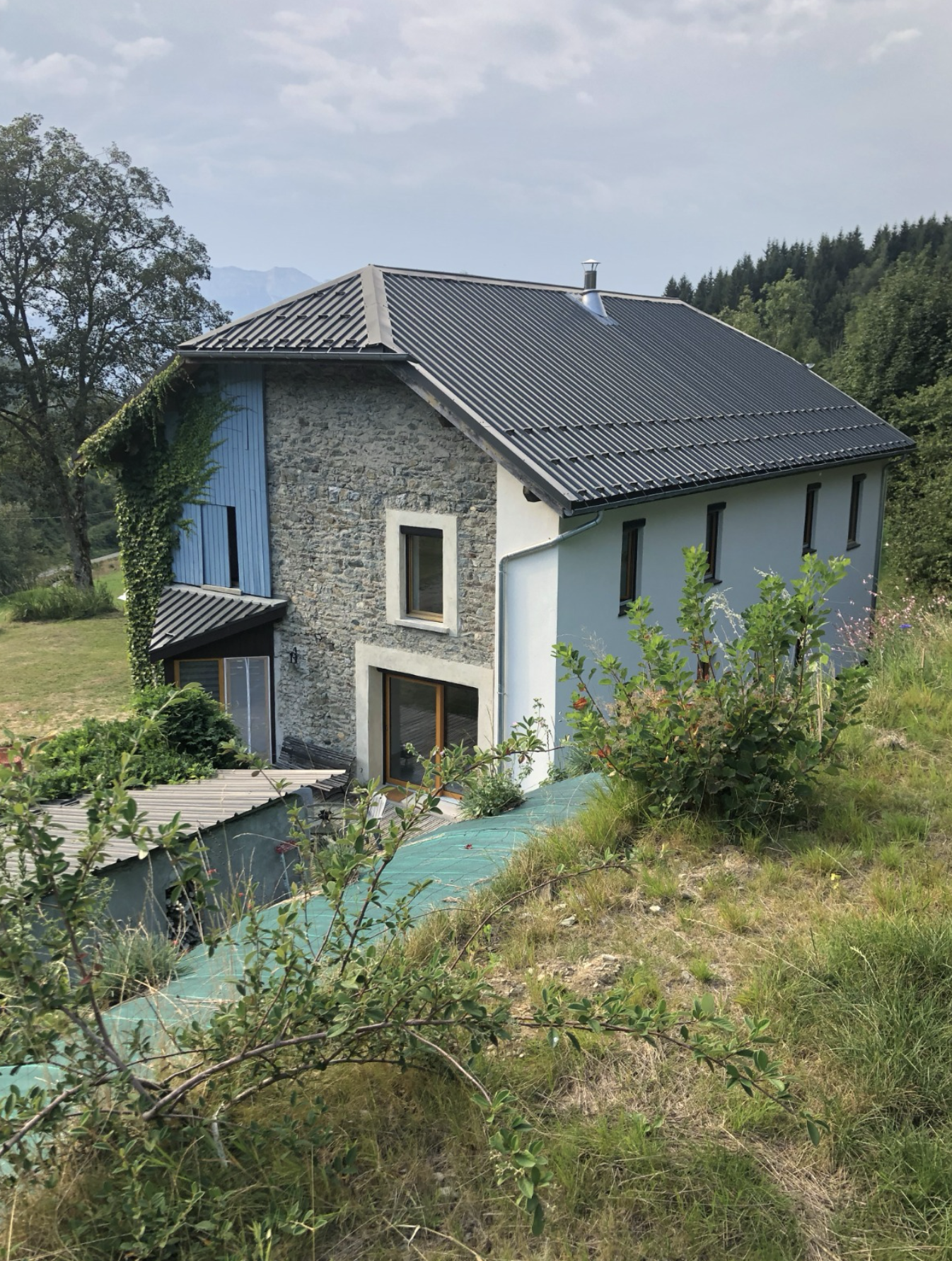 A house with a stone section and white walls, black window frames, and aged black roofing in a green landscape with trees and hills.