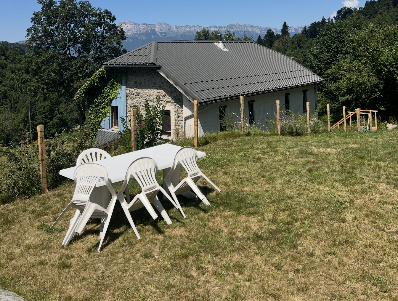 A grassy backyard with a white plastic table and four white plastic chairs, a stone house with a metal roof, surrounded by trees and mountains in the distance.