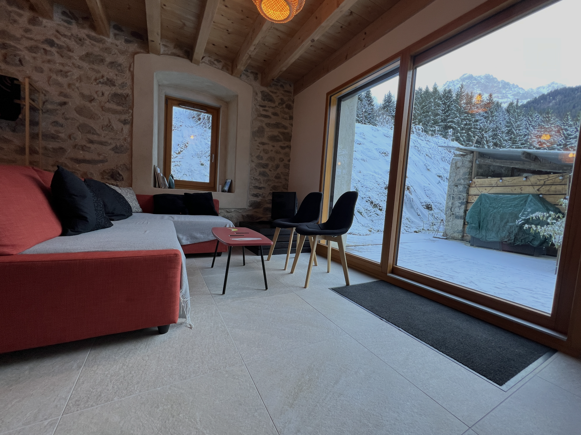 Interior of a cozy living room with large glass windows showing snowy outdoor mountains and trees. There is a red sofa with pillows, a small pink side table, and black chairs, with wooden ceiling beams and a stone wall.