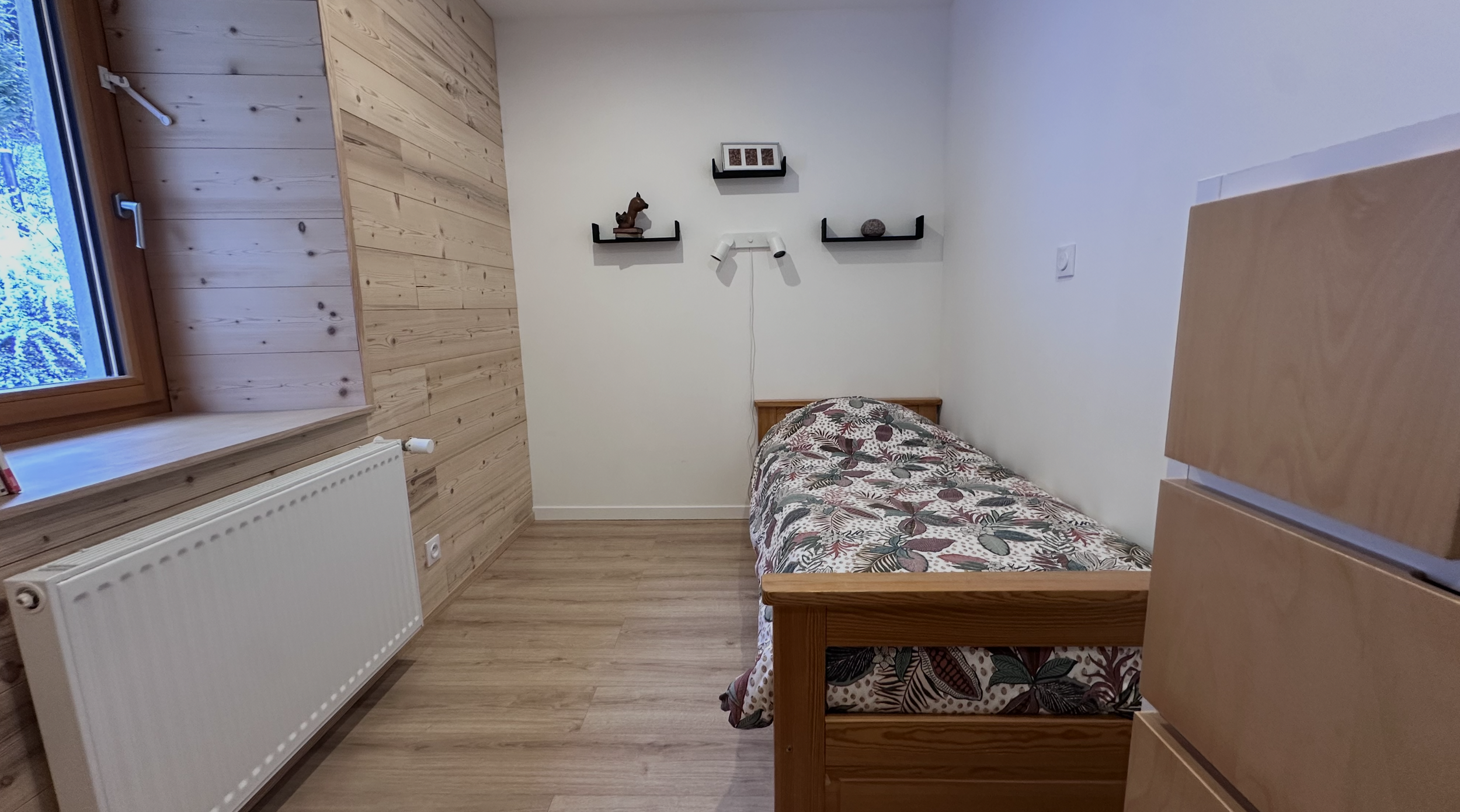 A small bedroom with light wood floors and paneling on one wall. There is a window with a snowy landscape outside. The room has a single bed with a floral patterned blanket, a white radiator, and a wall with three small black shelves holding decorative items.