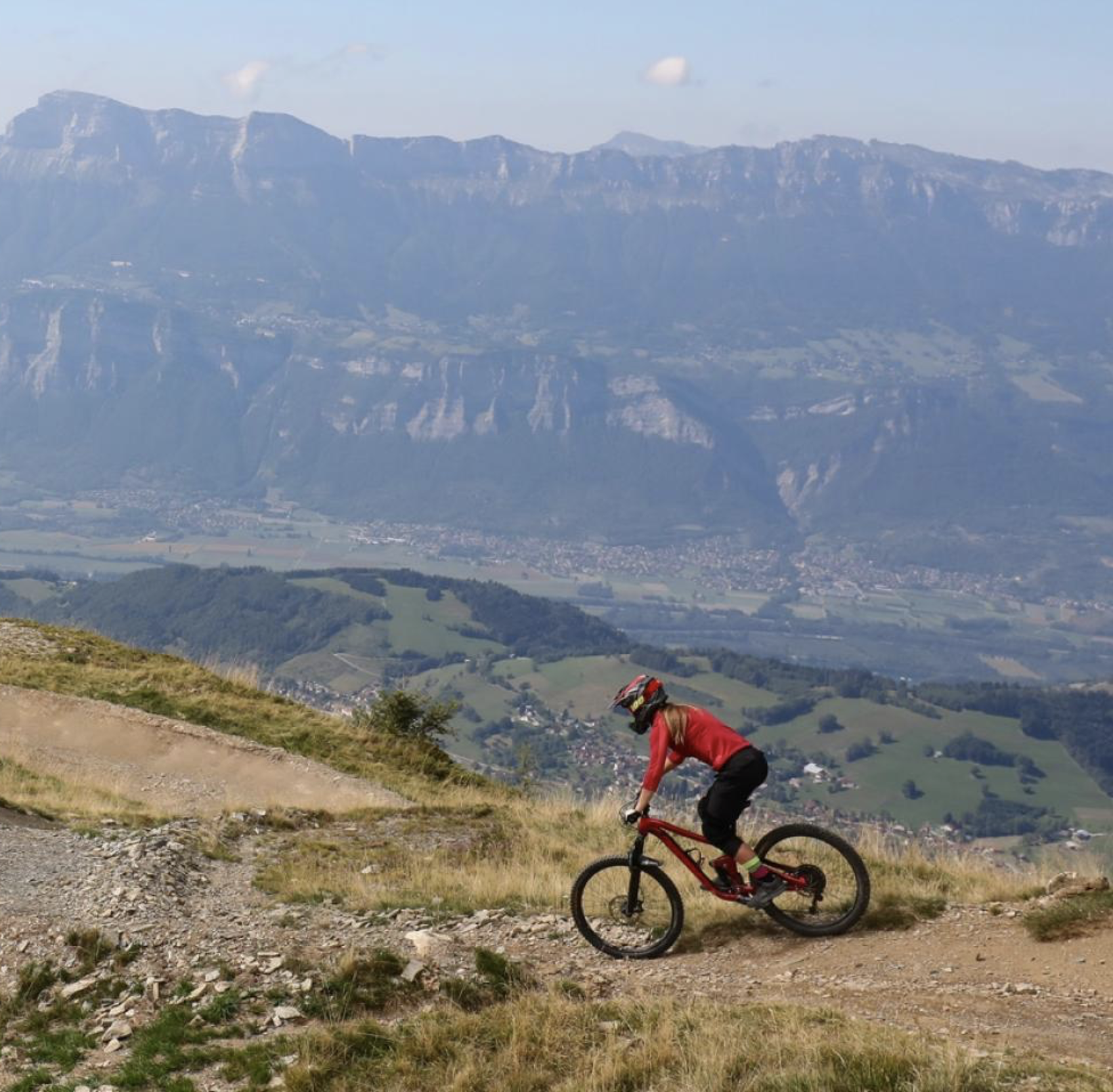 A person wearing a red jacket and a helmet riding a mountain bike on a dirt trail in a mountainous landscape with hills and a town in the distance.