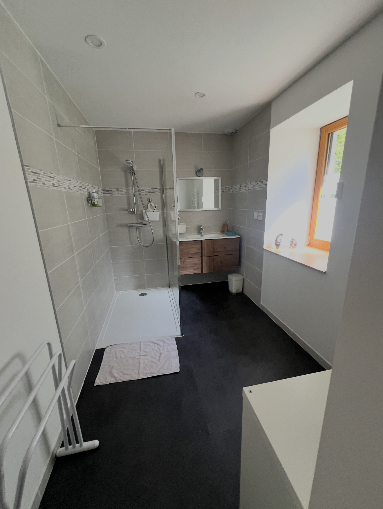 A modern bathroom featuring a walk-in shower with a glass panel, beige tiled walls with decorative border, a wooden vanity with a sink, a mirror, a window letting in natural light, and a dark floor with a white bath mat.