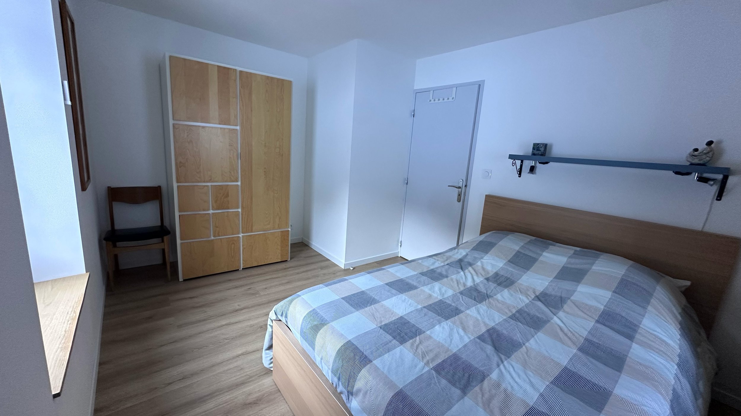 A bedroom with a bed, a wooden wardrobe, a chair, and a shelf with a decorative item. The room has wooden flooring and white walls.