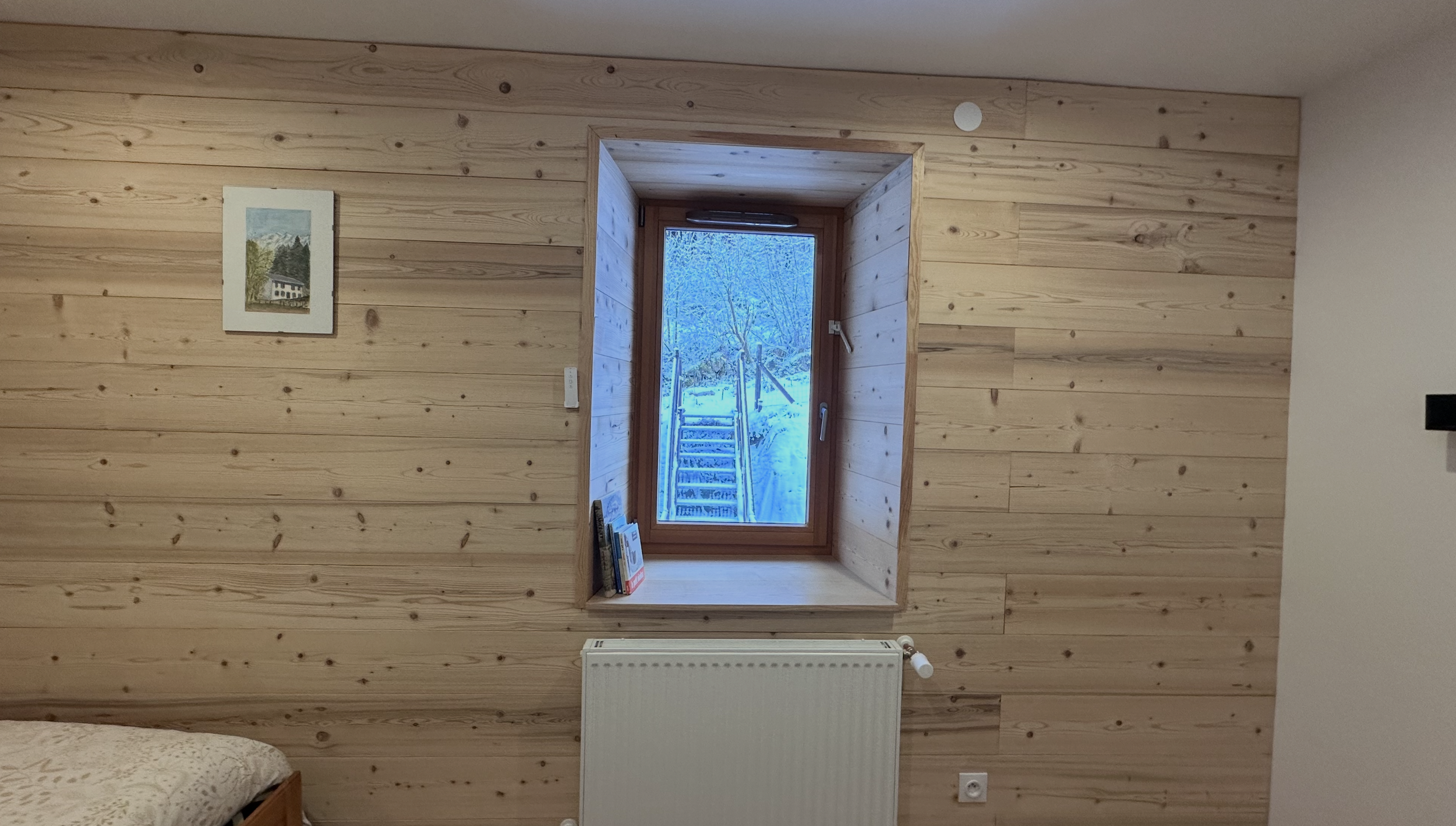 A room with wooden paneled walls, a window looking out to snowy trees and outdoors, a small bookshelf with books, a white radiator below the window, and a bed partially visible in the corner.