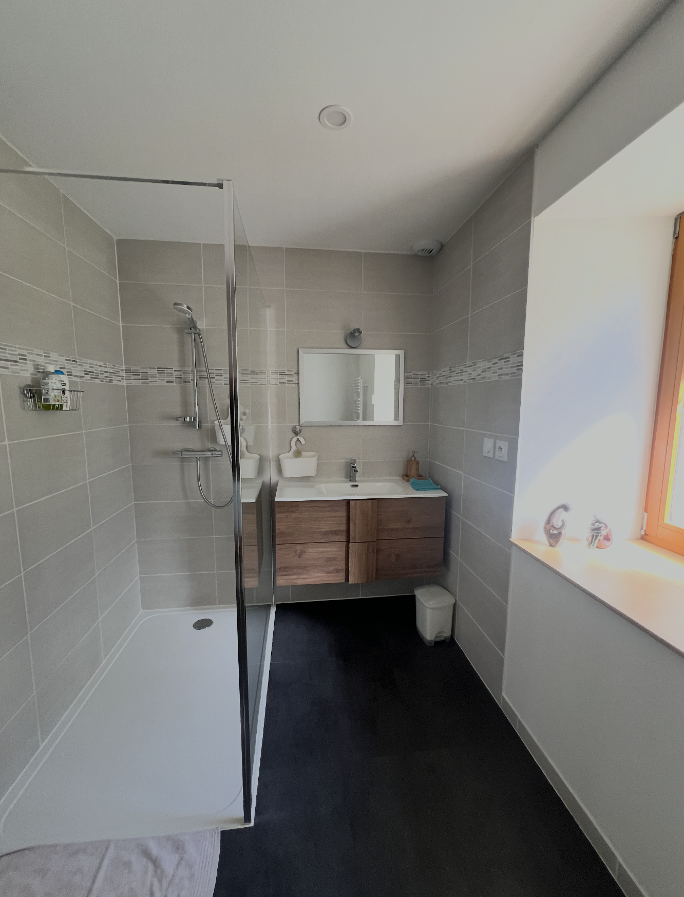 Modern bathroom featuring a walk-in shower with a glass door, beige tiled walls with a decorative strip, a wooden vanity with a mirror, a small sink, and a window allowing natural light.