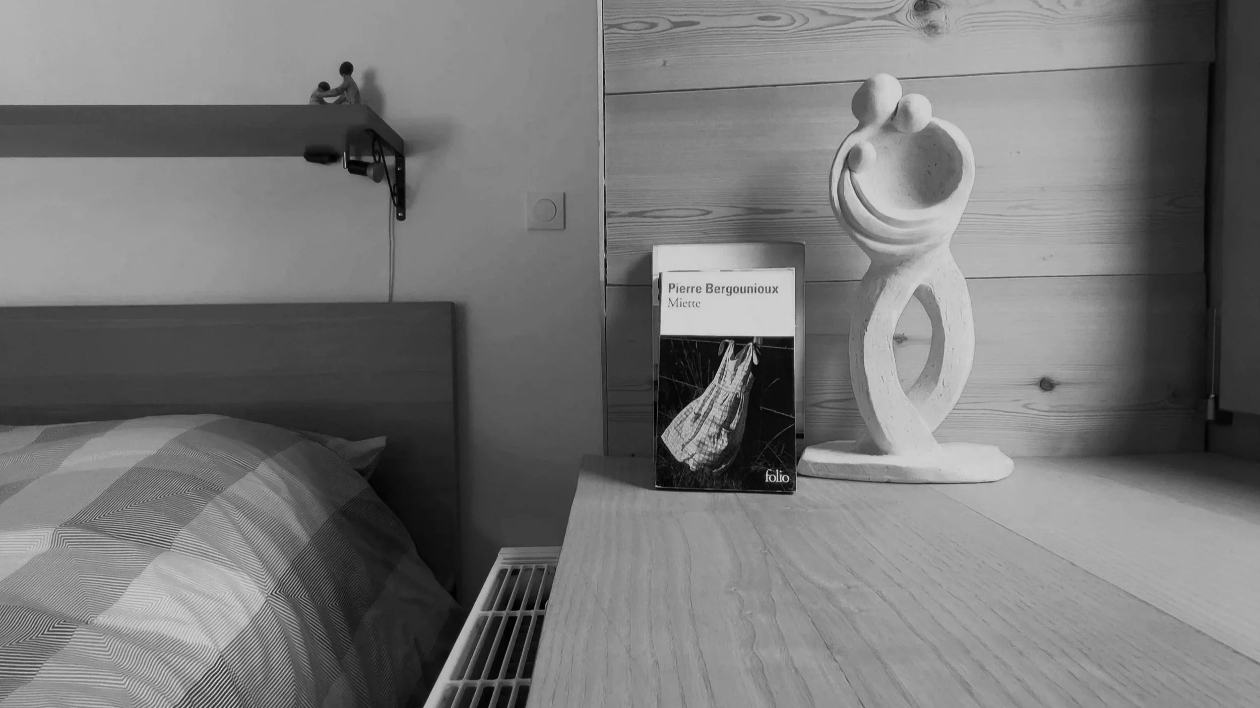 A bedroom corner with a wooden headboard, a bedside table, a sculpture resembling a simplified human figure, a book titled 'Pierre Bergounioux Miette', and a decorative shelf with two small figurines.