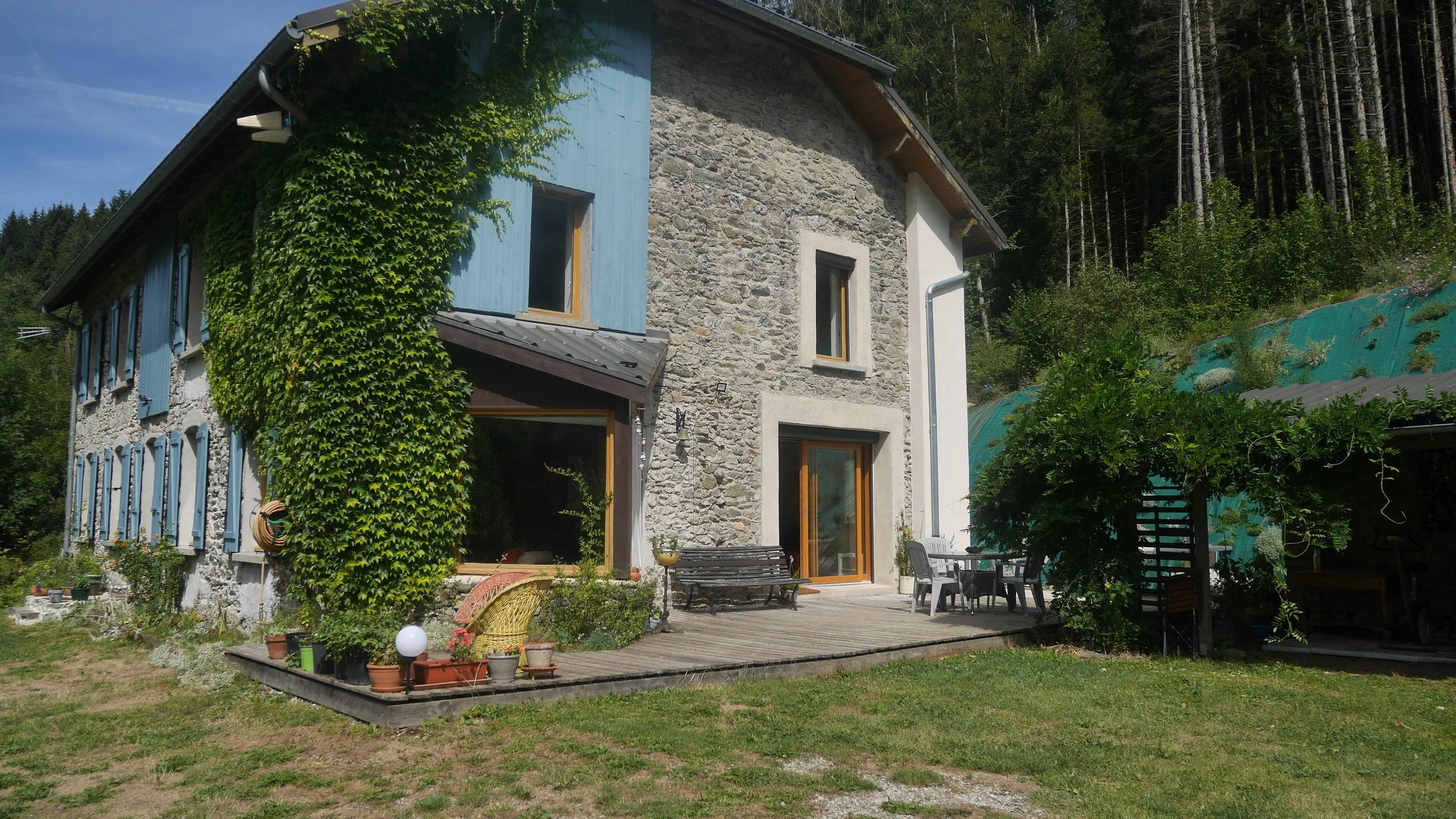 A two-story house with a stone and painted exterior, surrounded by a garden and outdoor furniture. The house has windows with blue shutters, some covered in ivy. There is a wooden patio with a table, chairs, and a bench, and a background of forested hills under a blue sky.