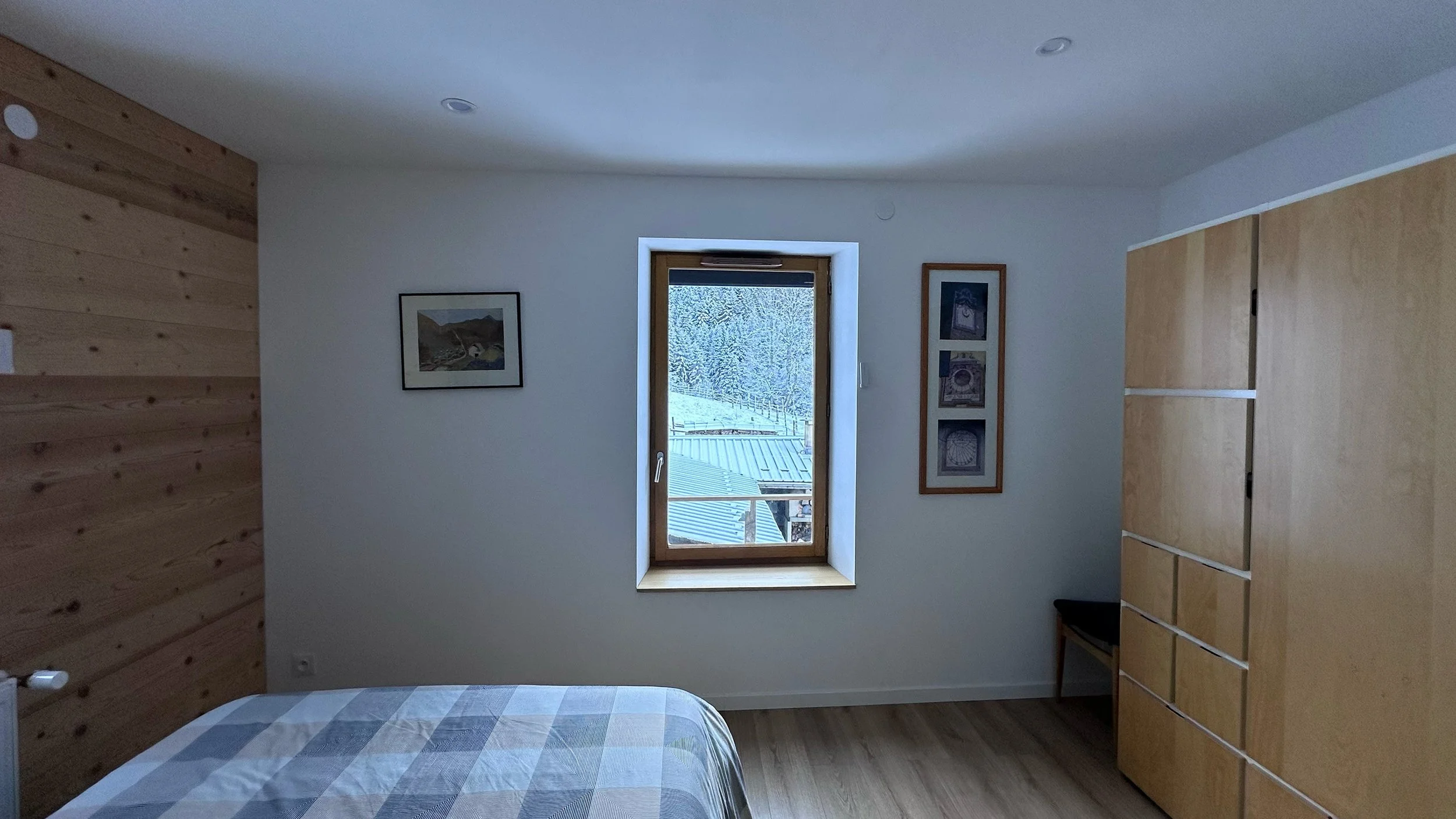 A bedroom with a window showing a snowy landscape, framed artwork on the wall, wooden furniture, and a bed with a checkered blanket.