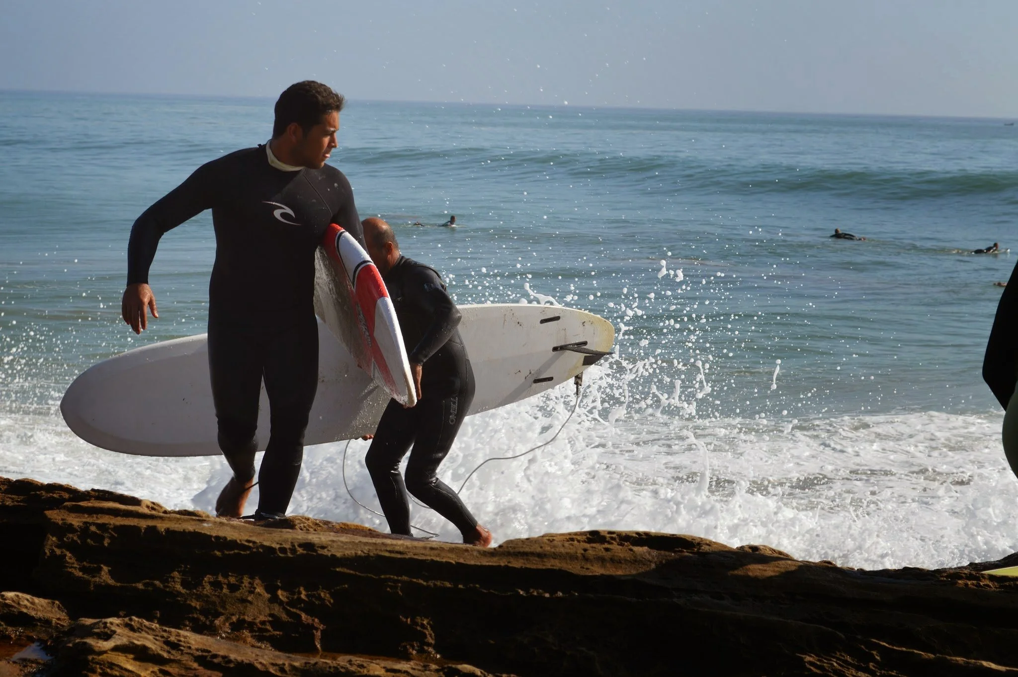 People in wetsuits carrying surfboards onto the beach near the water, with other surfers in the ocean and waves in the background.
