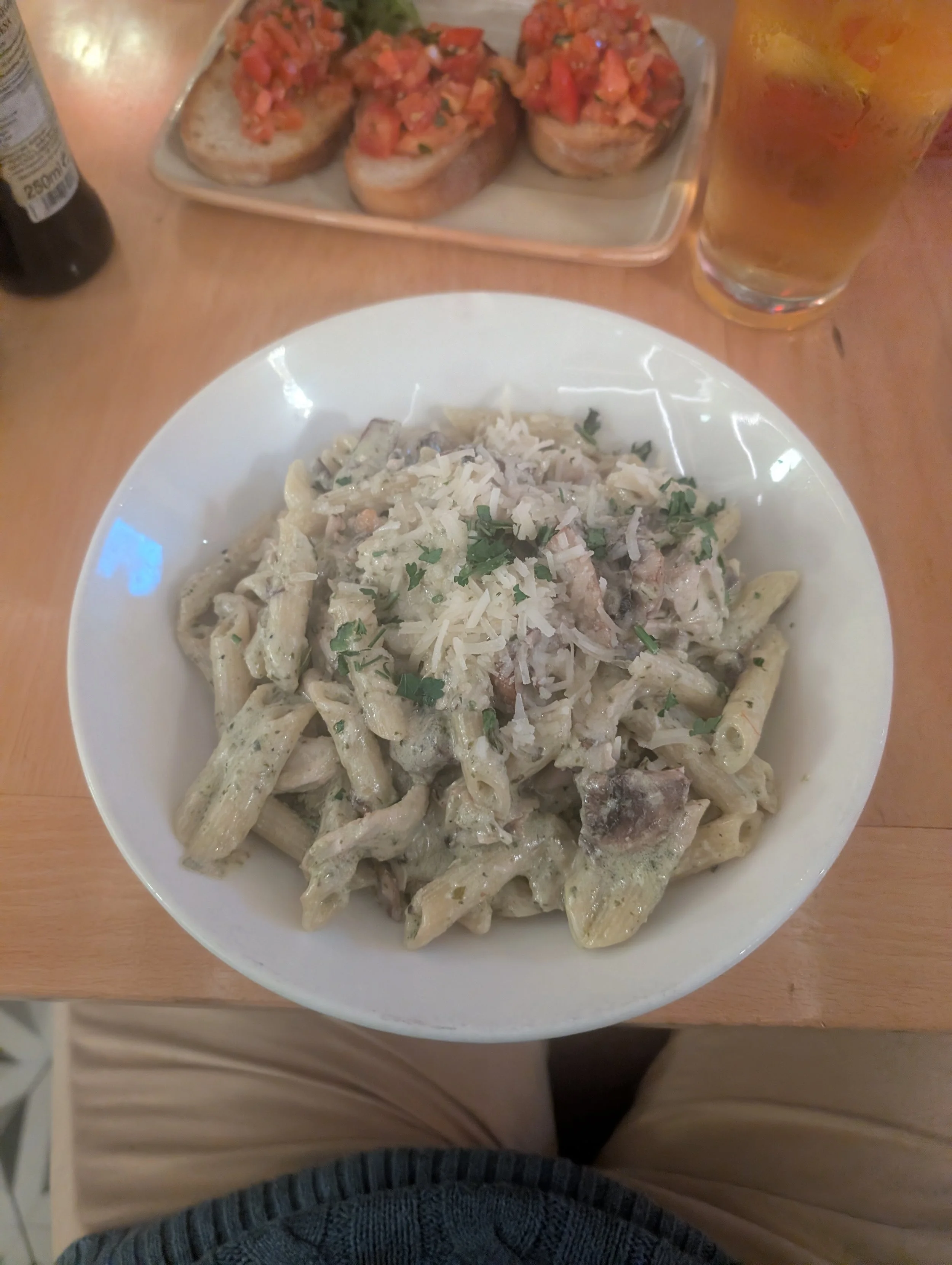 A plate of creamy pasta with herbs, a bowl of tomato bruschetta on toasted bread, and a glass of iced tea on a wooden table.