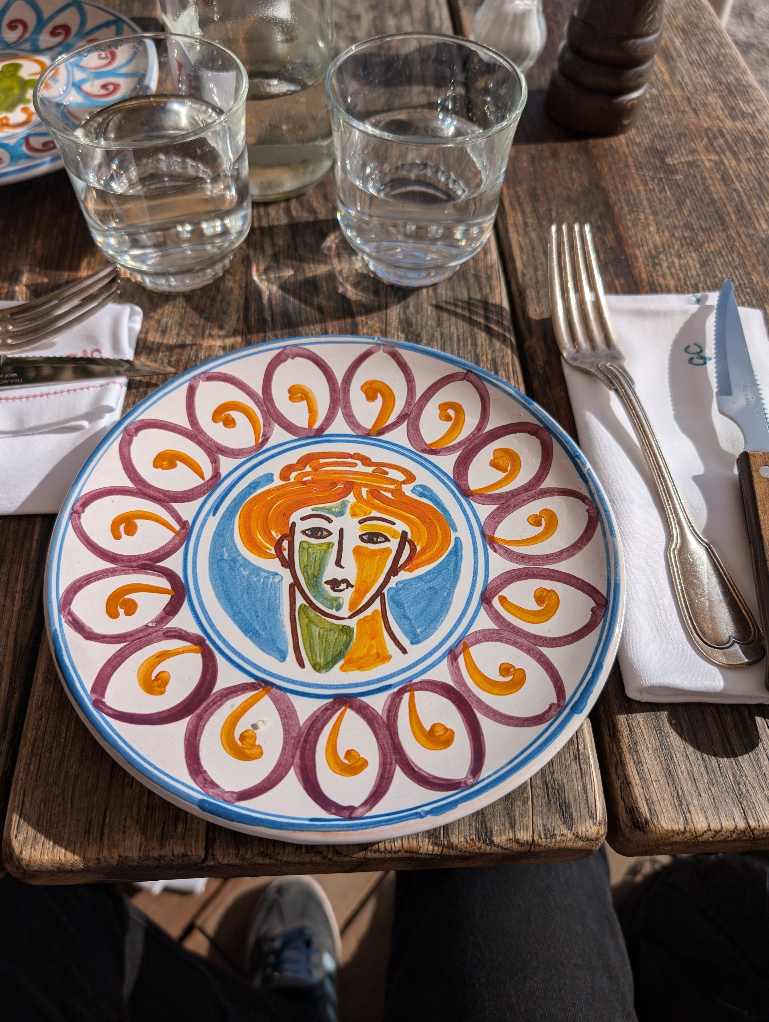 A colorful decorative plate with a stylized face and flower pattern, placed on a rustic wooden table. Two glasses of water and a fork and knife are arranged on a white napkin next to the plate.