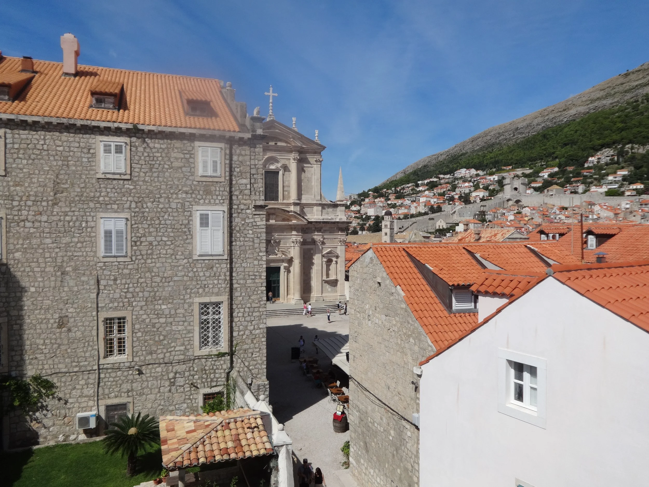 View of historic stone buildings with orange tiled roofs, a courtyard with people, and a hillside with white houses in the background under a blue sky.