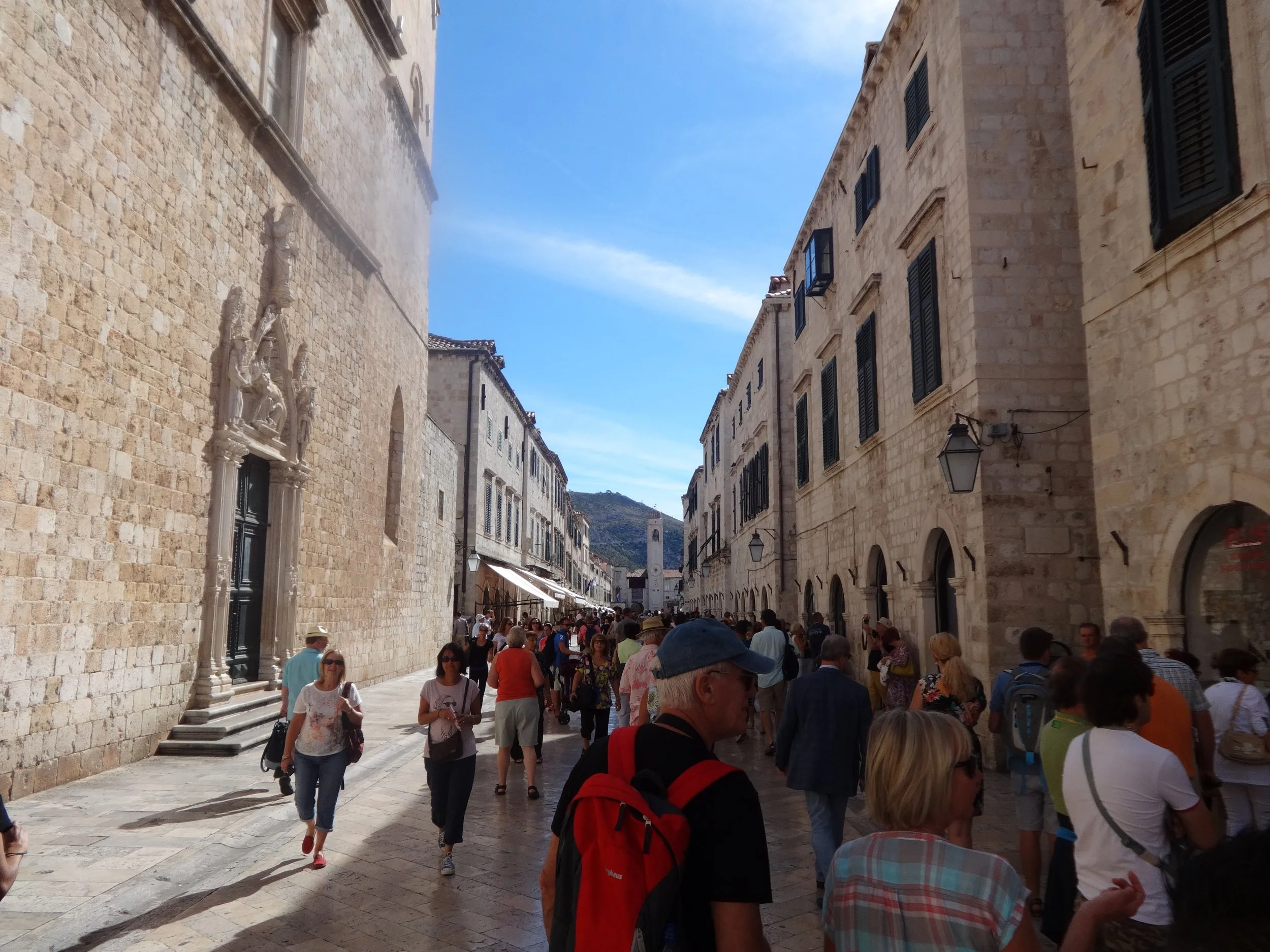 Crowded street scene in a historic European city with stone buildings, black shutters, and a visible clock tower in the background. People are walking, shopping, and sightseeing under a clear blue sky.