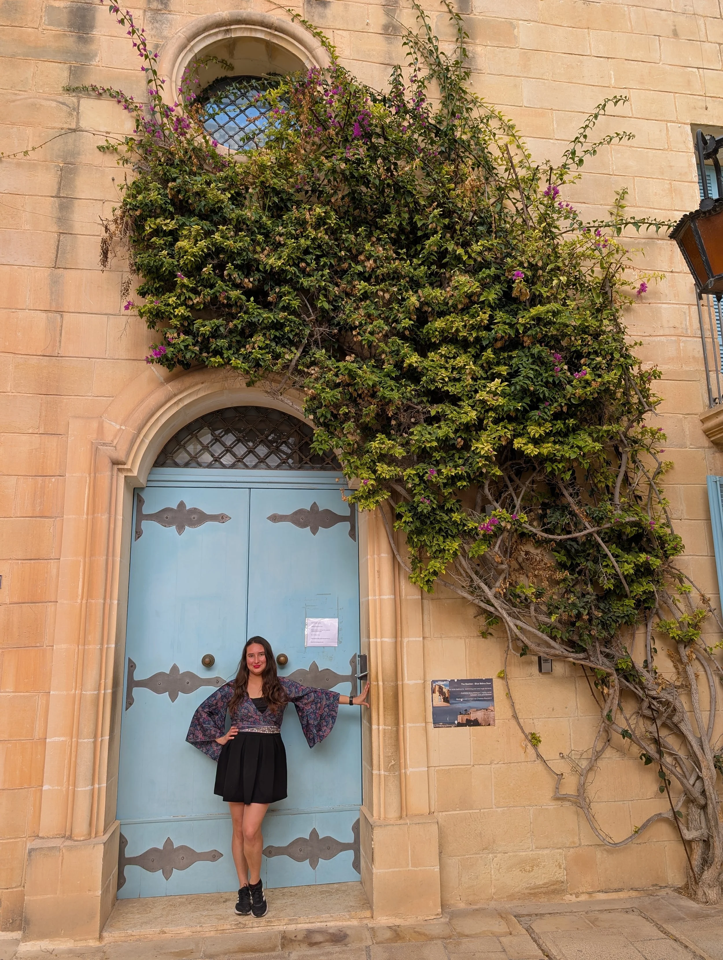 A woman with long dark hair wearing a dark floral top and a black skirt standing in front of a large turquoise door with black ironwork, framed by a beige stone wall. She has her left hand on her hip and her right hand holding the door handle, smilin