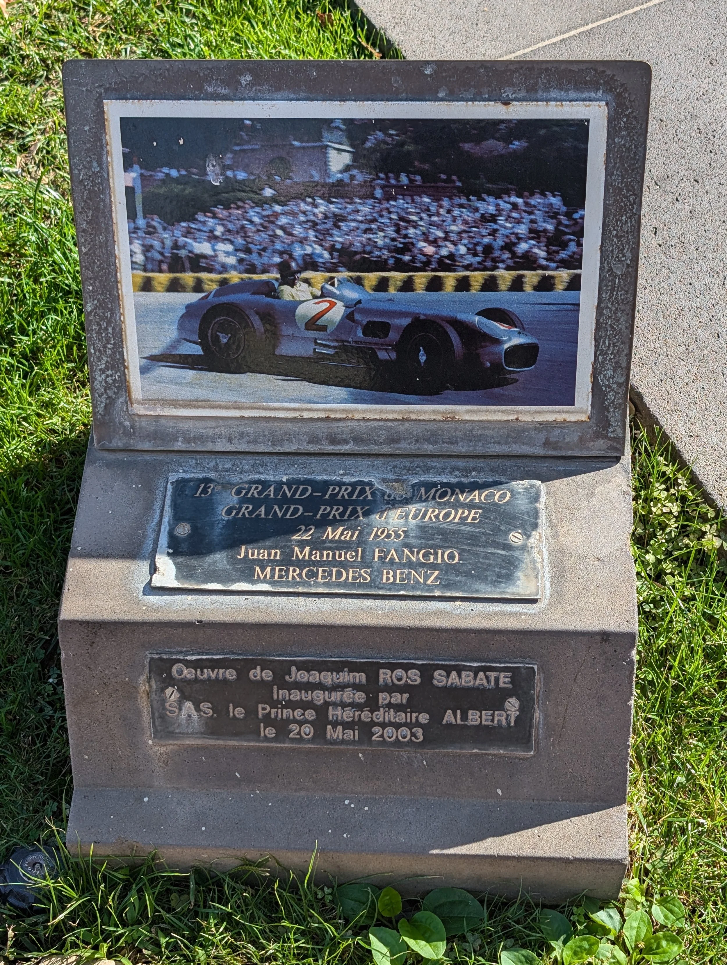 A commemorative plaque featuring a color photo of a vintage race car with driver, and text in French about the 1955 Monaco and Europe Grand Prix, with additional French text about an inauguration in 2003.
