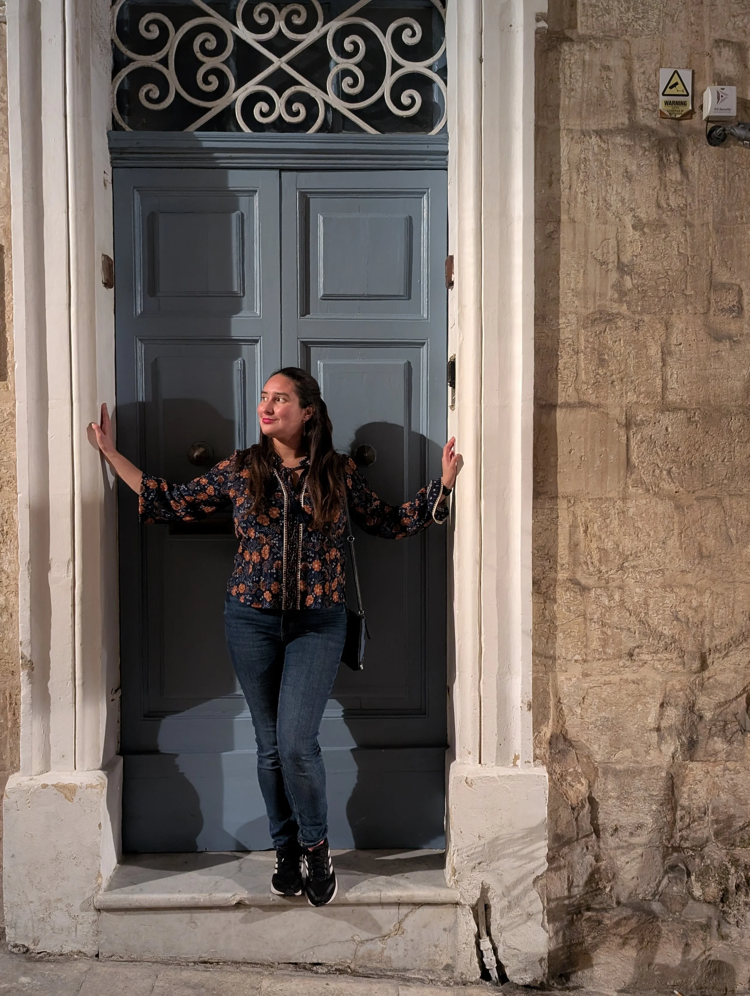 A woman with long dark hair, wearing a dark floral blouse and jeans, standing in front of a large blue door with her hands touching the door frame. The door and the woman are set in a stone wall with a small security camera and warning signs on the u