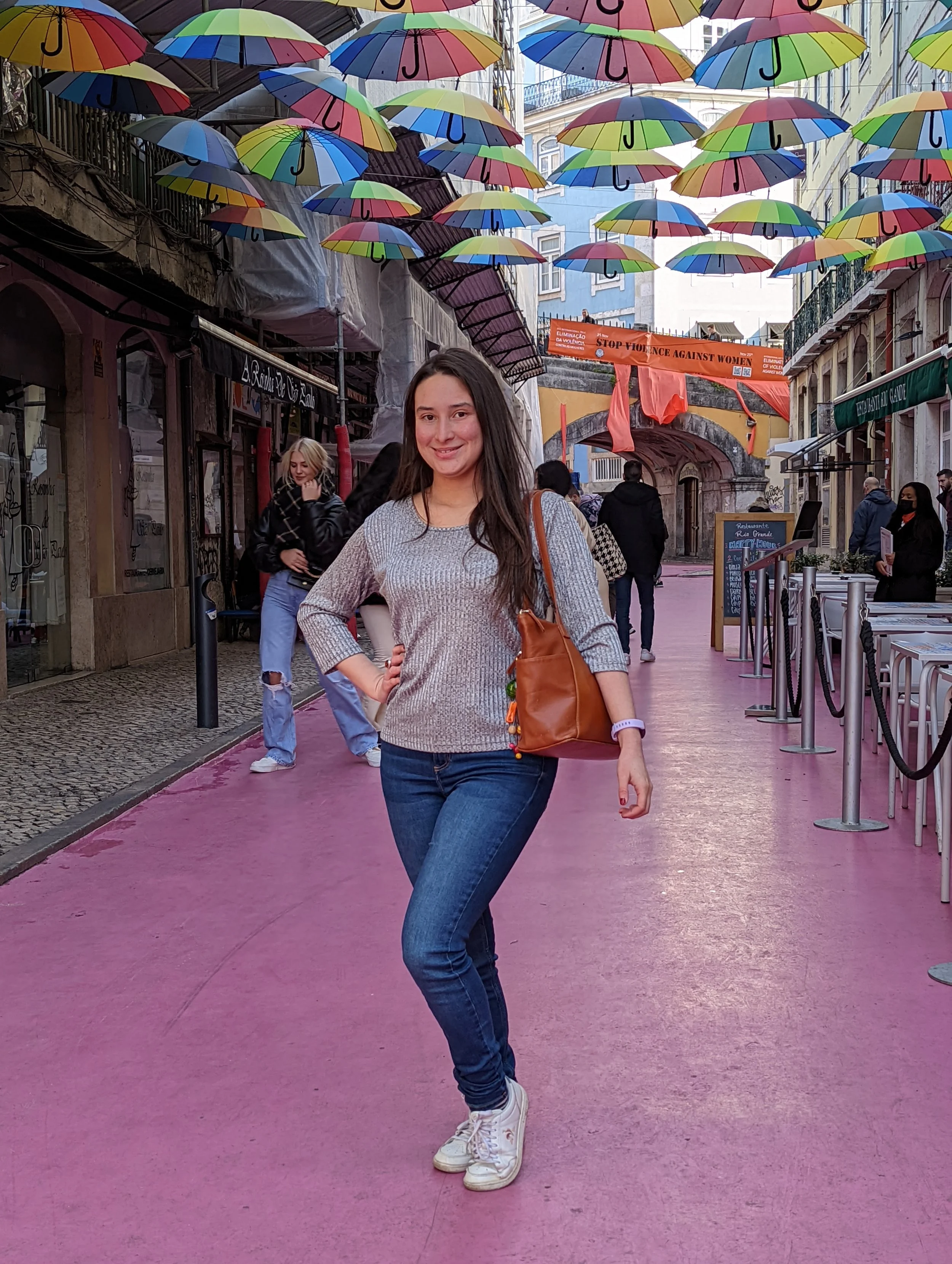 Young woman standing on a pink walkway under colorful umbrellas in an urban street with shops and other people in the background.
