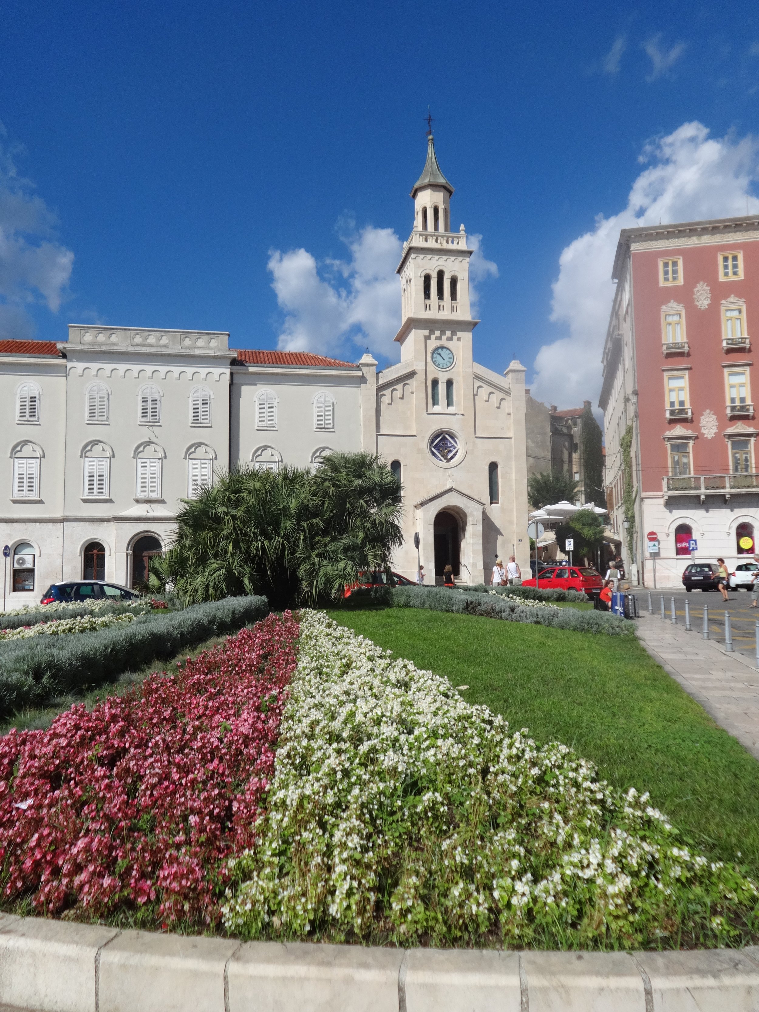 A city square with a historic church, colorful buildings, a flower bed with pink and white flowers, and a clear blue sky.