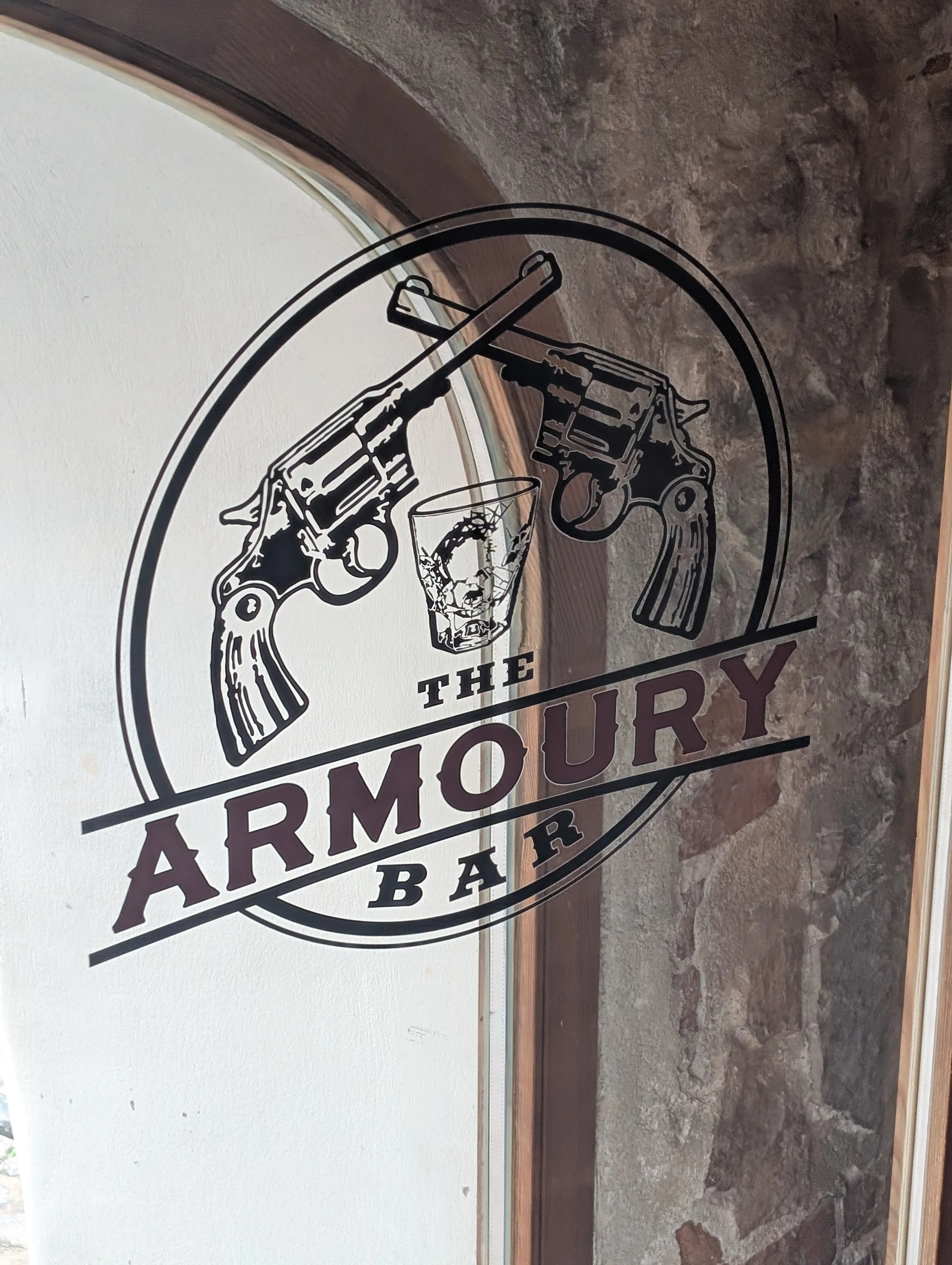 A metal sign with a revolver and a handgun pointing toward a whiskey glass, with the words "THE ARMORY BAR" and "BAR".
