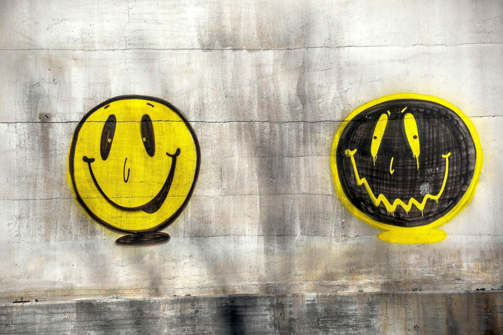 Two smiley face graffiti art on a concrete wall, one bright yellow with a happy expression, and the other black with a wavy smile and an ominous expression, both outlined in yellow.