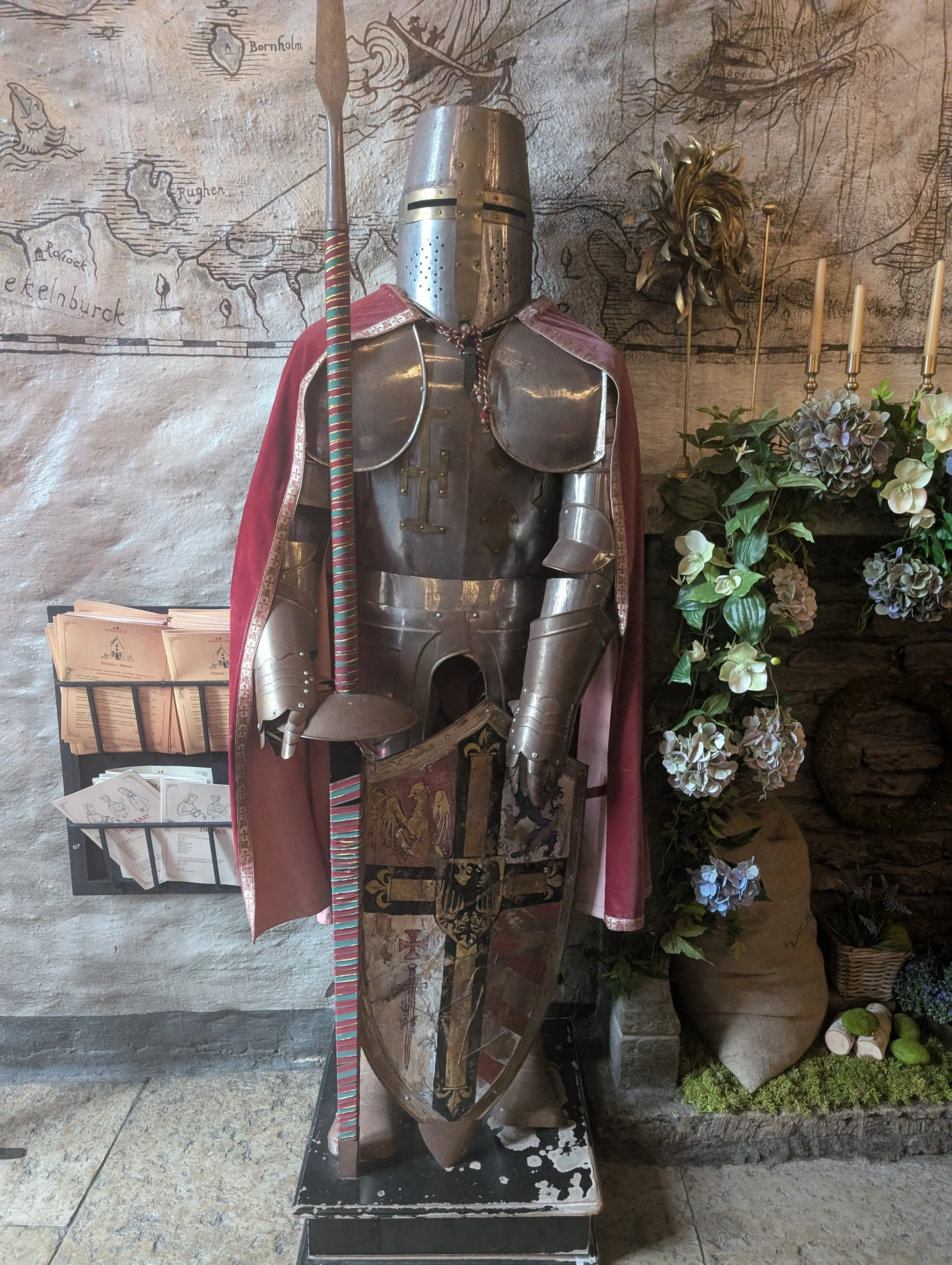 A suit of medieval armor with a helmet, chest plate, and gauntlets, draped in a red cape with gold trim. The armor is displayed against a wall with a historic map or mural, and there is a figurine of a knight on horseback in the background. A staff w
