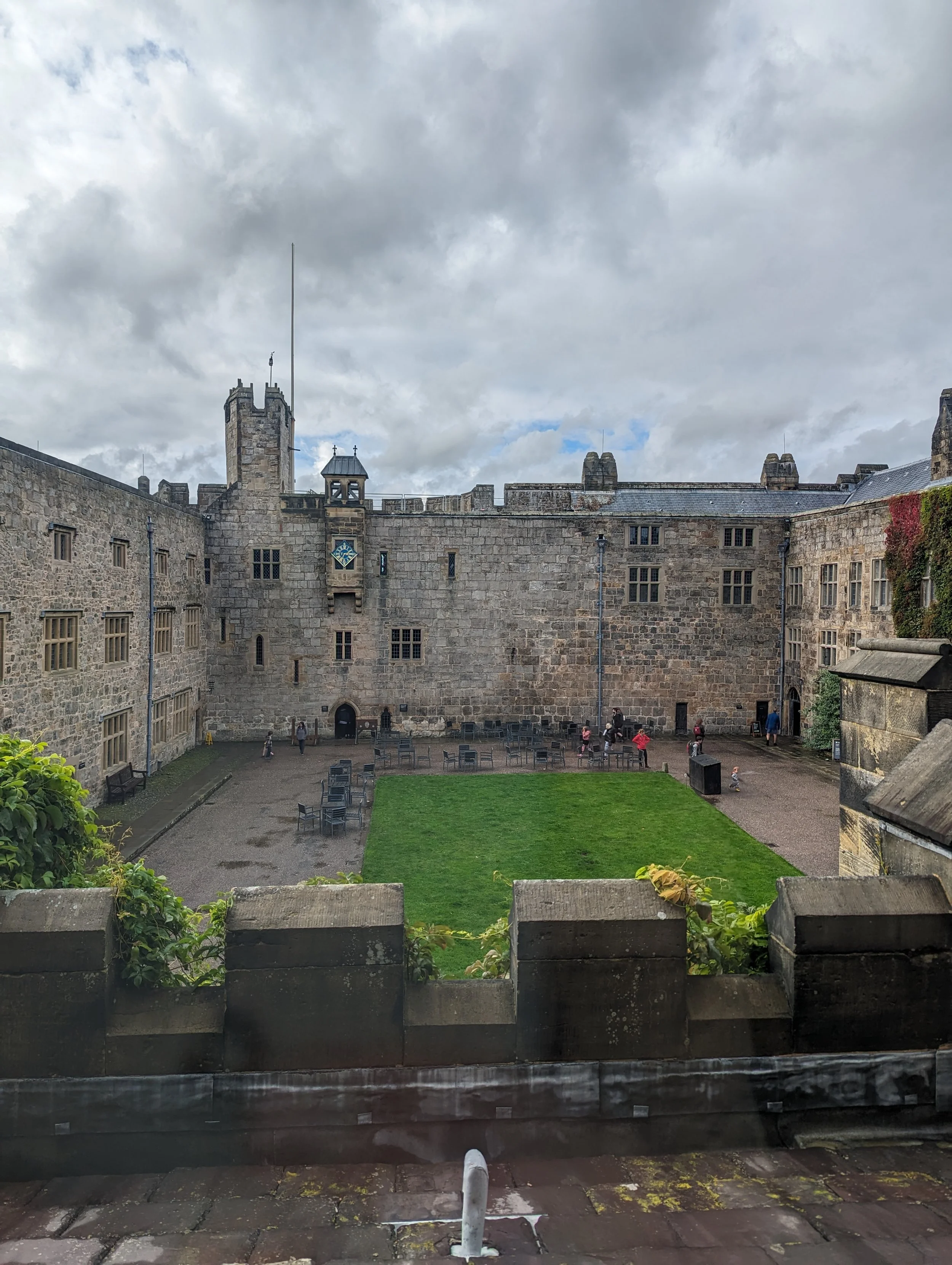 View of a historic stone castle courtyard with benches, green lawn, and cloudy sky.