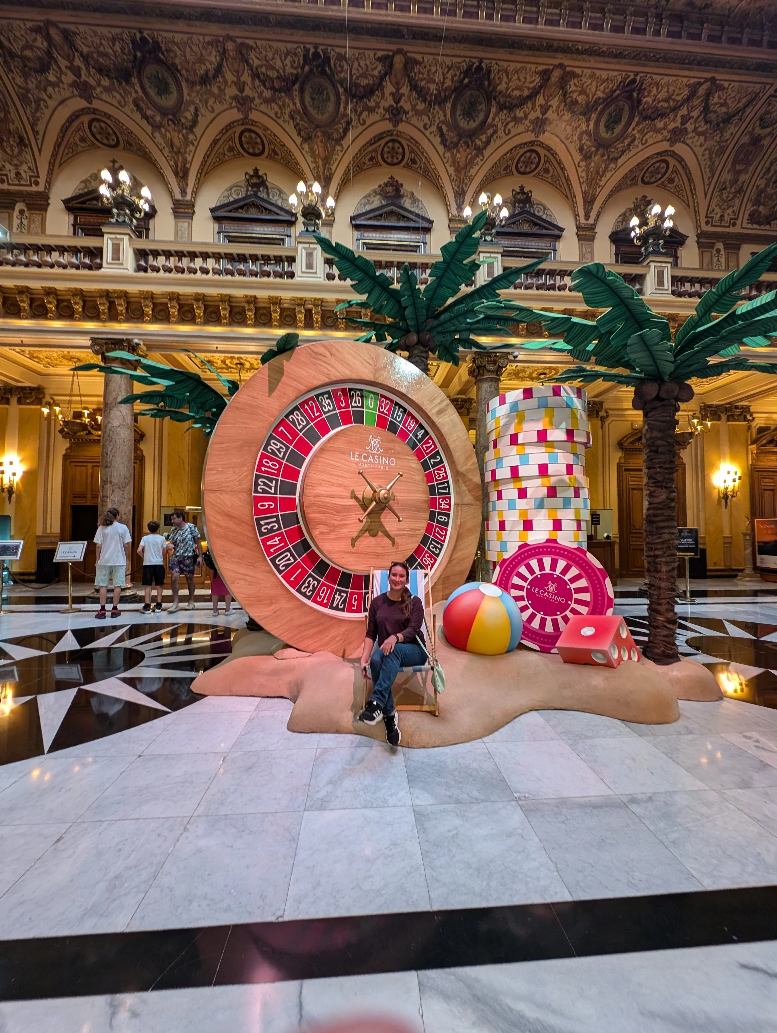 Interior of a grand hotel lobby with a roulette wheel themed display, two artificial palm trees, and colorful casino-themed props including oversized dice and a giant deck of cards. Several people are visible in the background.