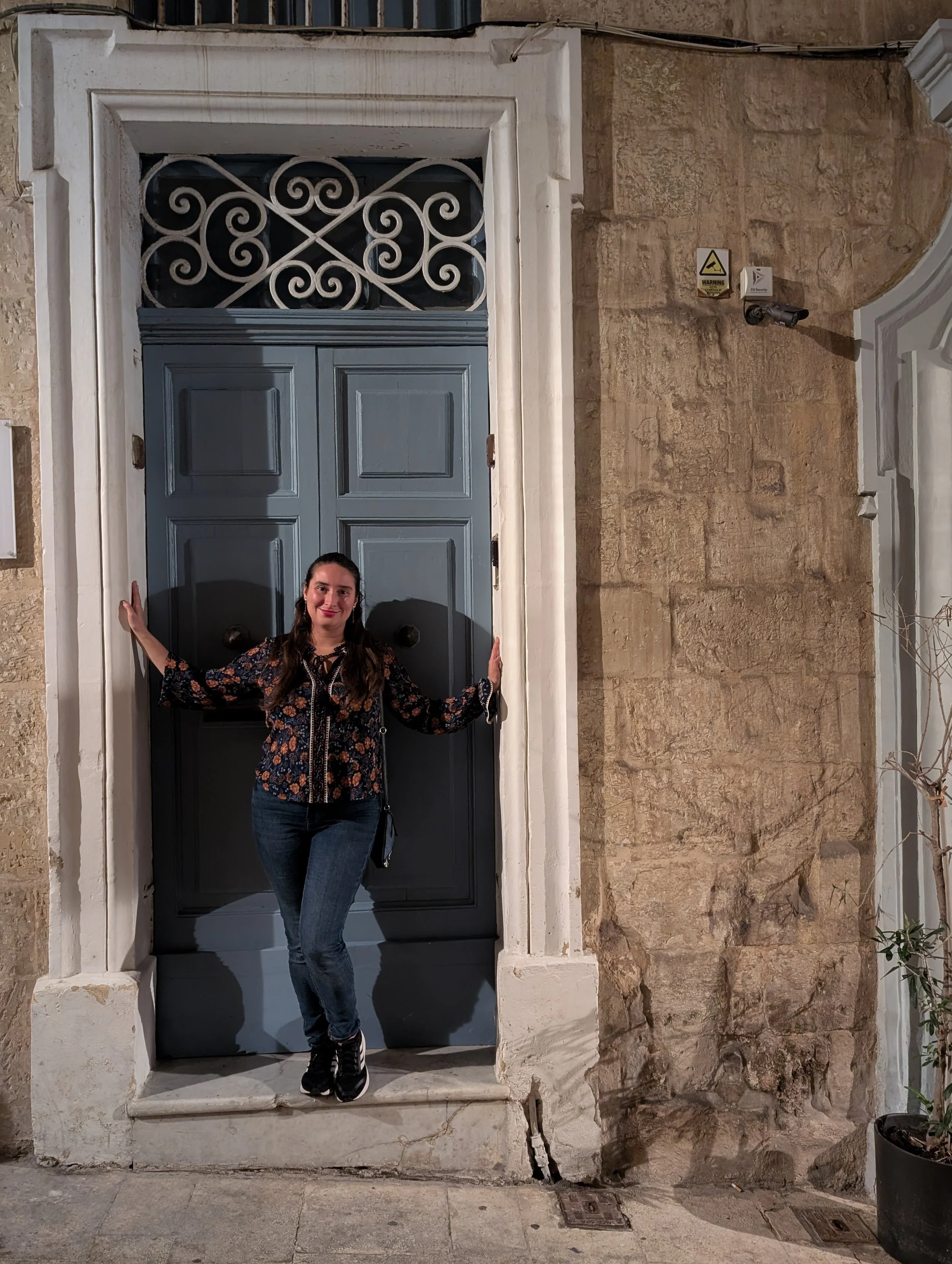 A woman standing in a doorway made of stone and wood at night, smiling, with arms outstretched touching the sides of the doorway. The background includes a textured stone wall and security cameras.