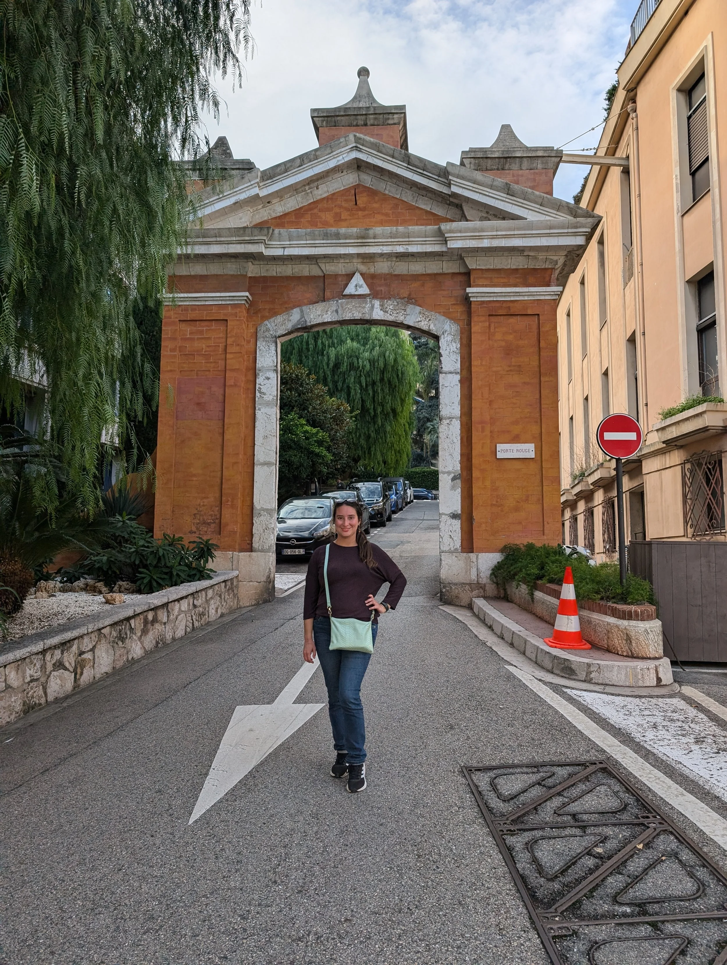 A woman standing on a city street in front of an archway with a historical building in the background. She is smiling and wearing a dark long-sleeve shirt and jeans, with a light-colored bag over her shoulder. There are parked cars along the street a
