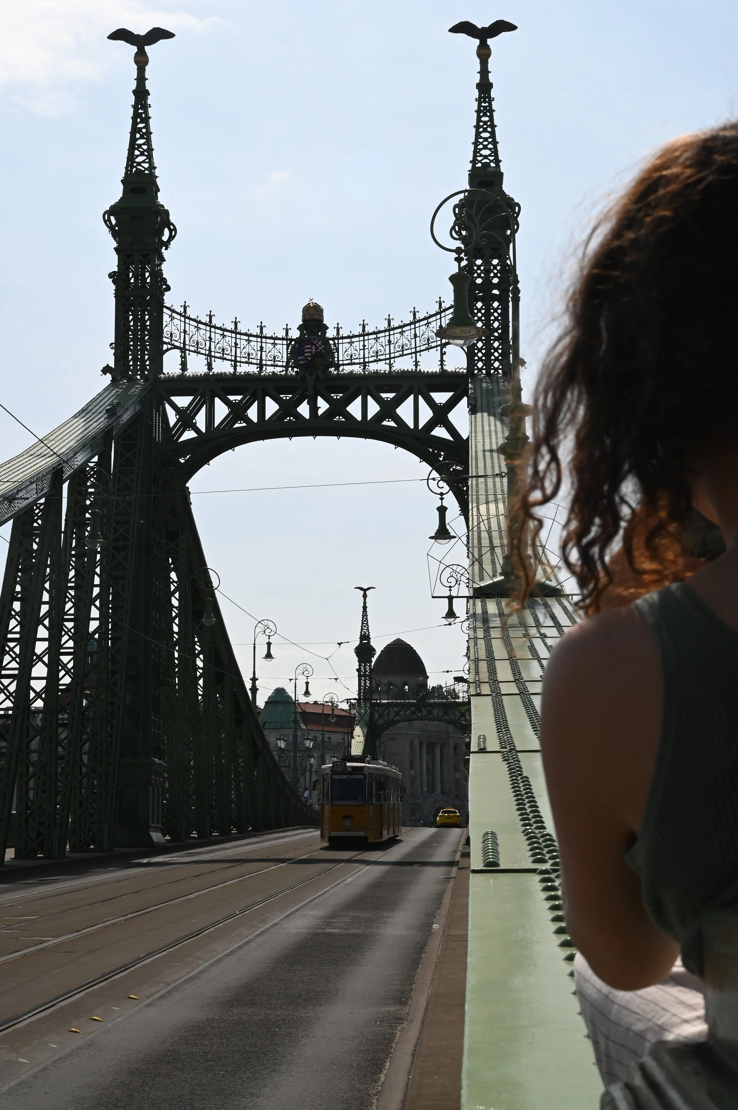 Street scene with a bridge featuring tall towers with eagle statues on top, tram tracks, and a tram. A woman with curly hair is partially visible on the right side.