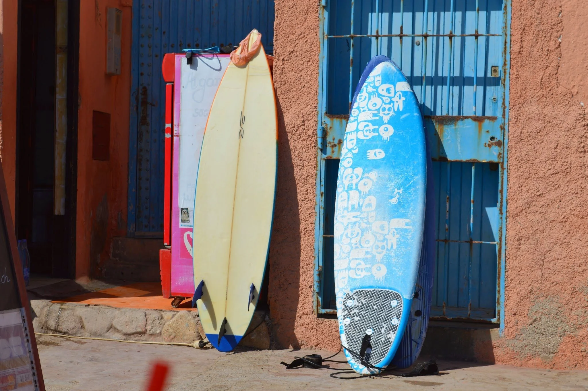 Colorful surfboards leaning against a rustic orange wall and blue metal gate.