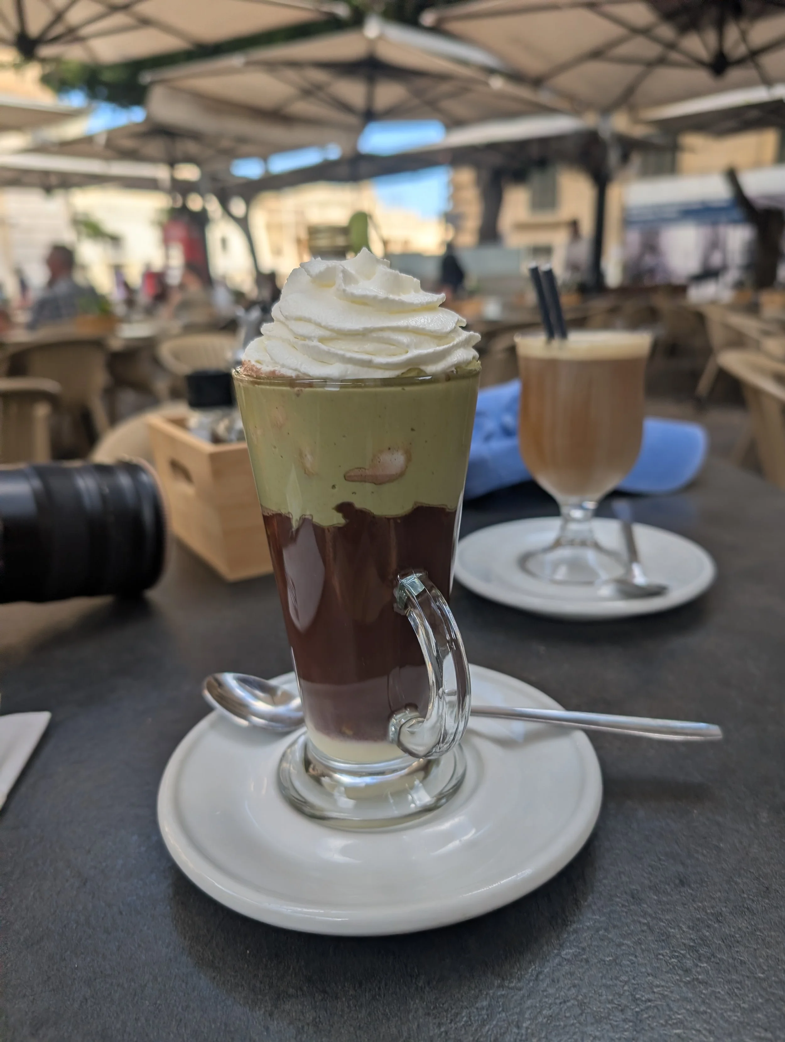 A tall glass of layered dessert with whipped cream on top, served on a white tray at an outdoor café.