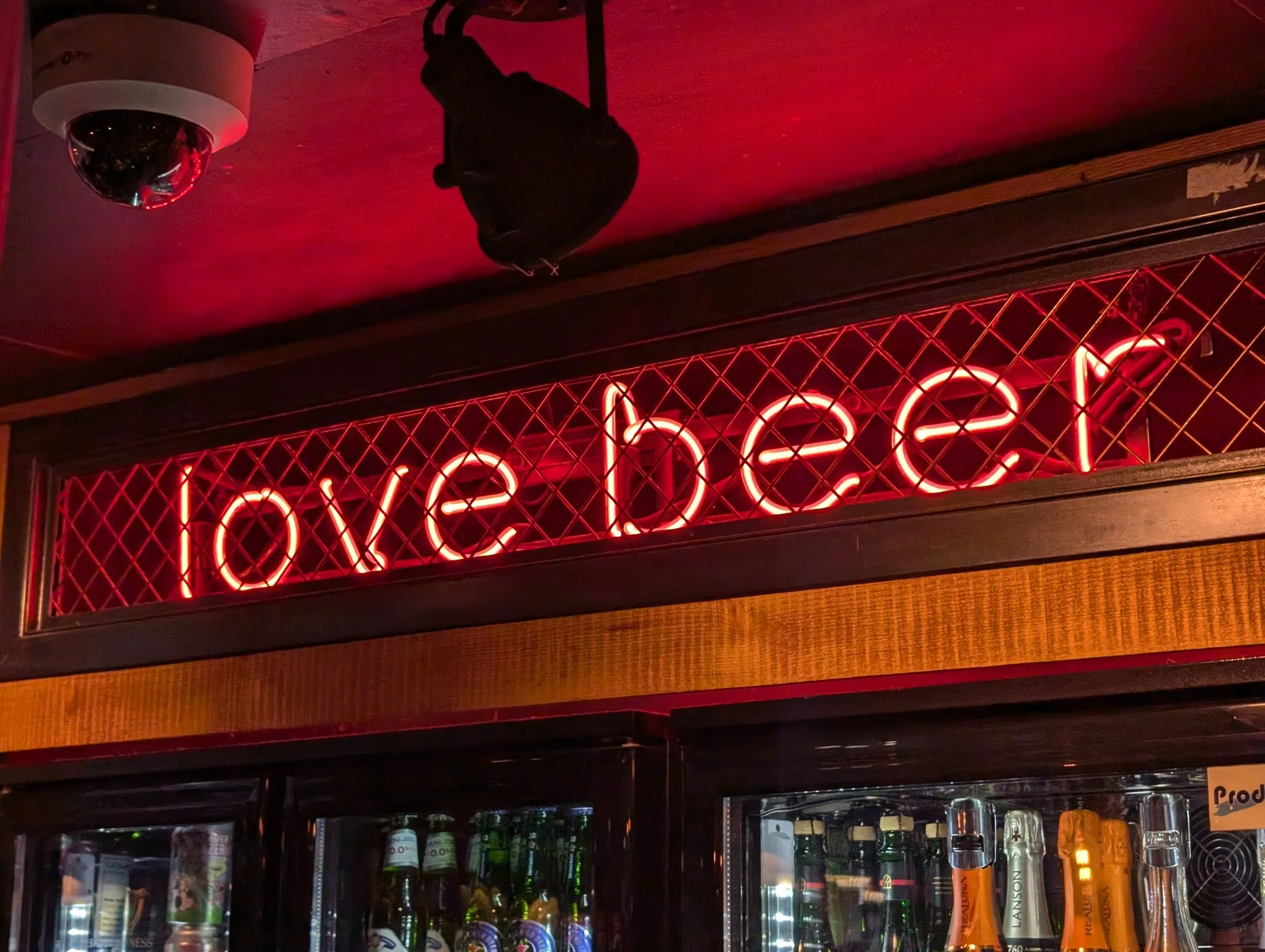 A neon red sign in front of a metal grid with the word 'OPEN' illuminated, hanging above a bar area with bottles and glasses visible behind the glass door of a refrigerator.