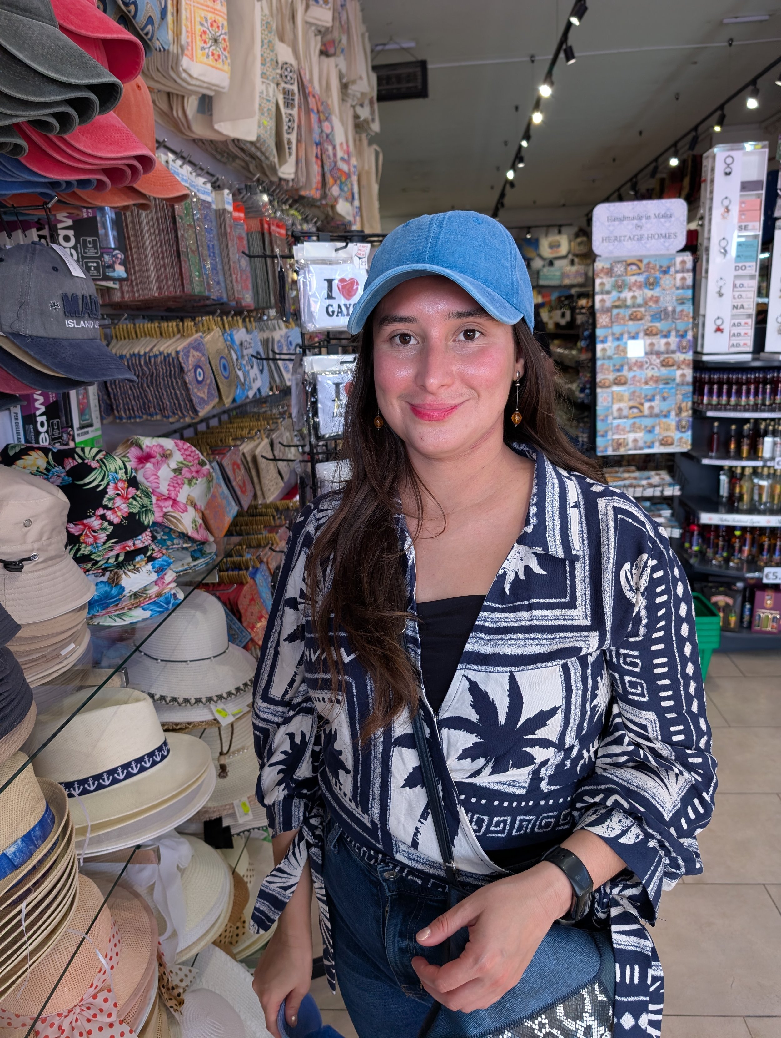 A woman with long brown hair, wearing a blue baseball cap, a navy blue shirt with a white floral pattern, and earrings, sitting in a store aisle with shelves of hats and souvenirs. She is smiling at the camera.