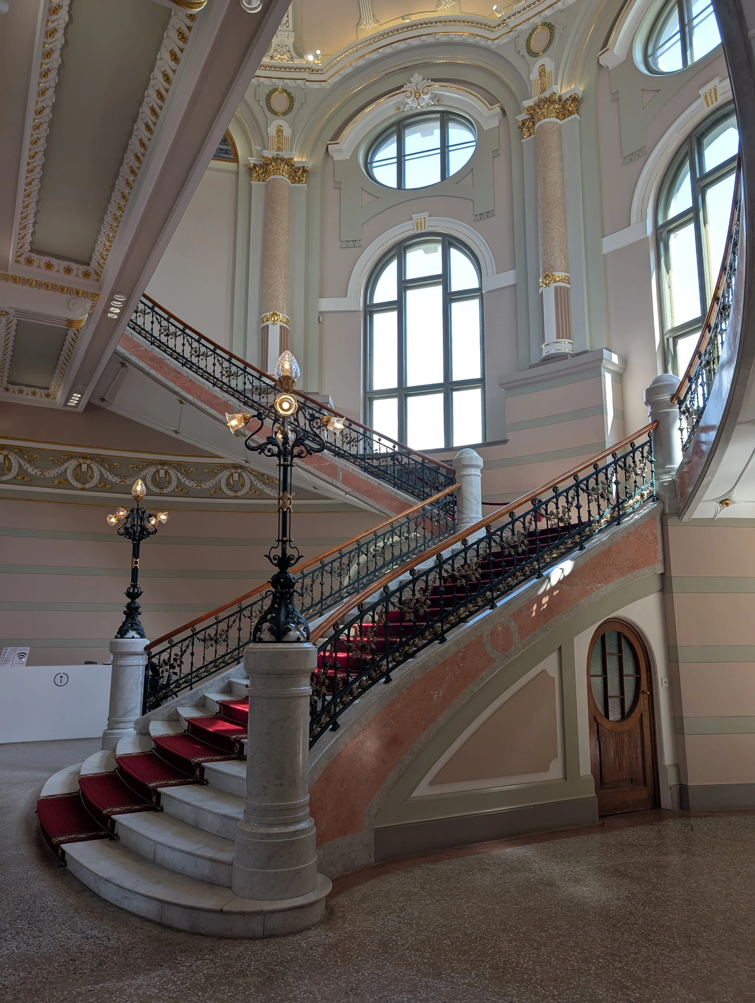 Interior view of a grand staircase with red carpet, ornate black iron railing, marble pillars with gold accents, large arched windows, decorative ceiling with gold and white details, and a chandelier.